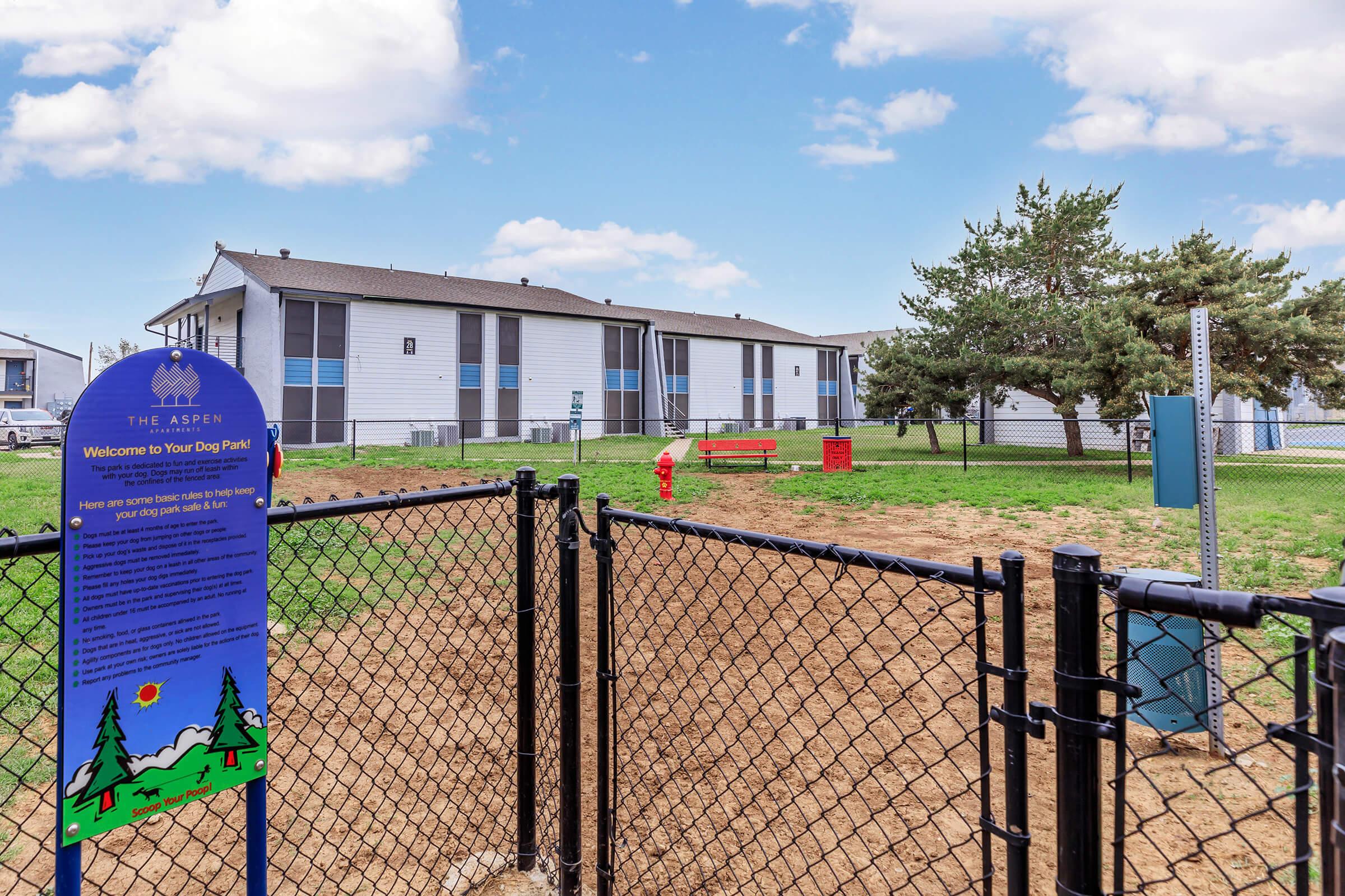 A fenced dog park area featuring an informational sign about the park, a bench, and trees in the background. Nearby, there are residential buildings under a blue sky with fluffy clouds. The ground is mostly grassy with some dirt patches, creating a welcoming space for pets and their owners.