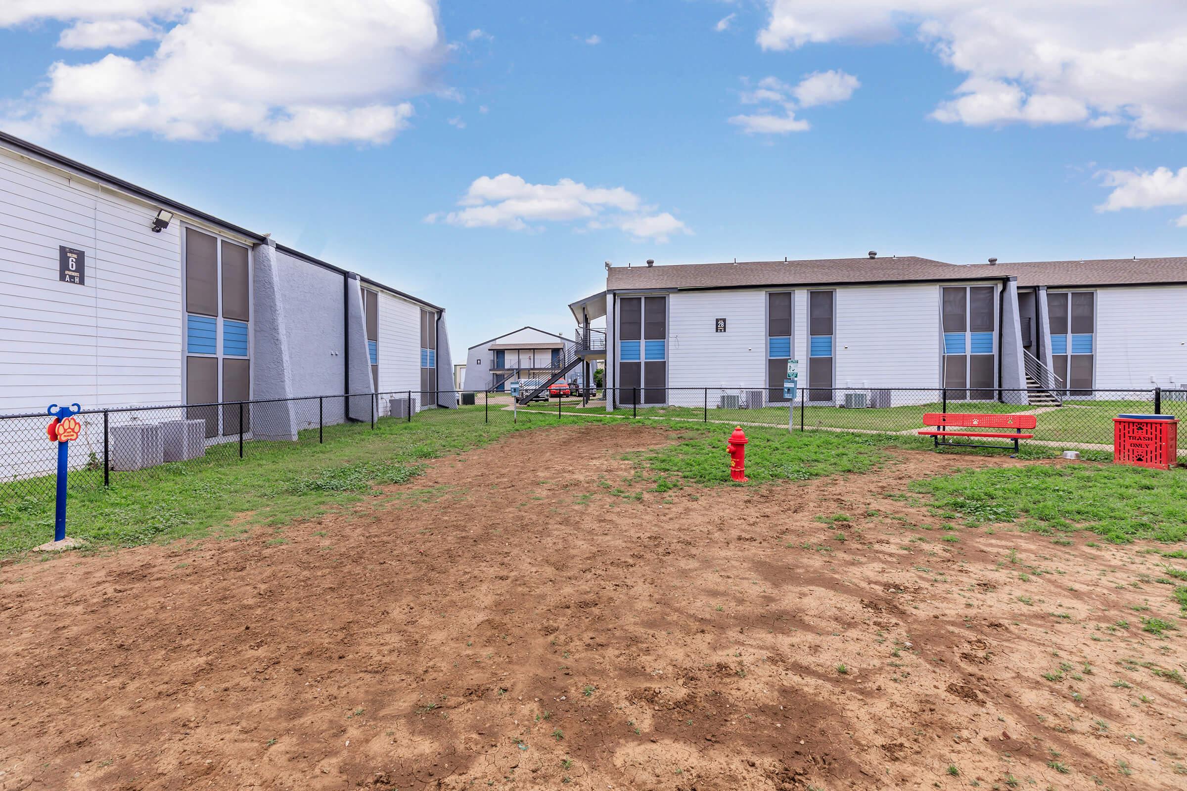 A dirt pathway leads between two multi-unit residential buildings in a grassy area. To the left, there's a blue sign with a swinging object, and a red bench sits beside a red fire hydrant. The sky is bright with scattered clouds.
