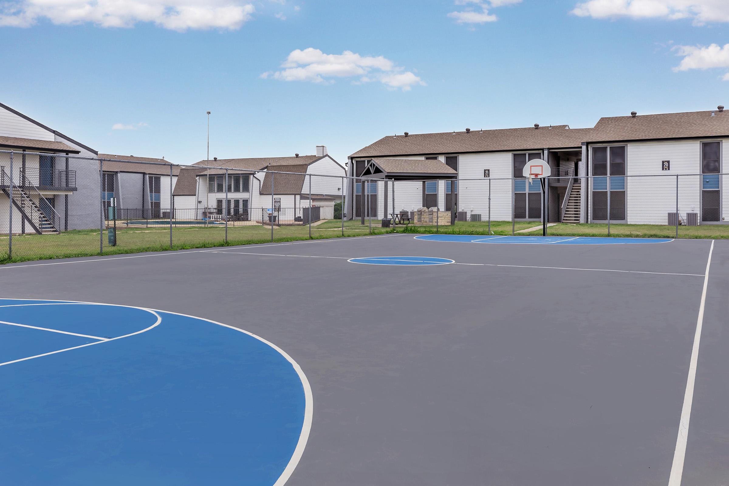 A basketball court featuring blue markings and a hoop, set against a backdrop of residential apartment buildings. Lush green grass surrounds the court, with clear blue skies overhead, creating a bright and inviting outdoor space.