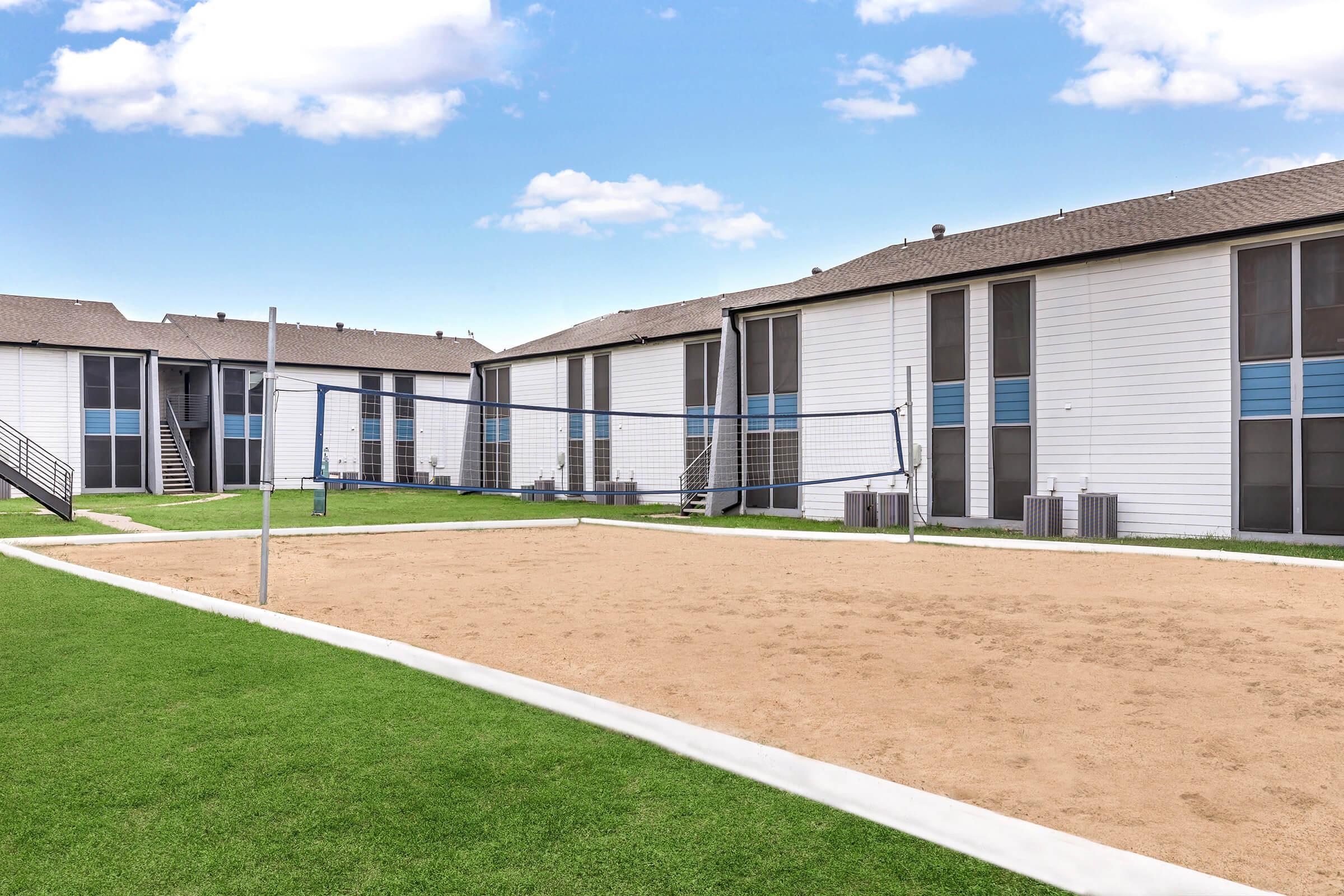 A sandy volleyball court surrounded by green grass, with two rows of white apartment buildings in the background. The buildings have large windows and a light exterior, and there are staircases leading to some entrances. The sky is blue with a few scattered clouds.