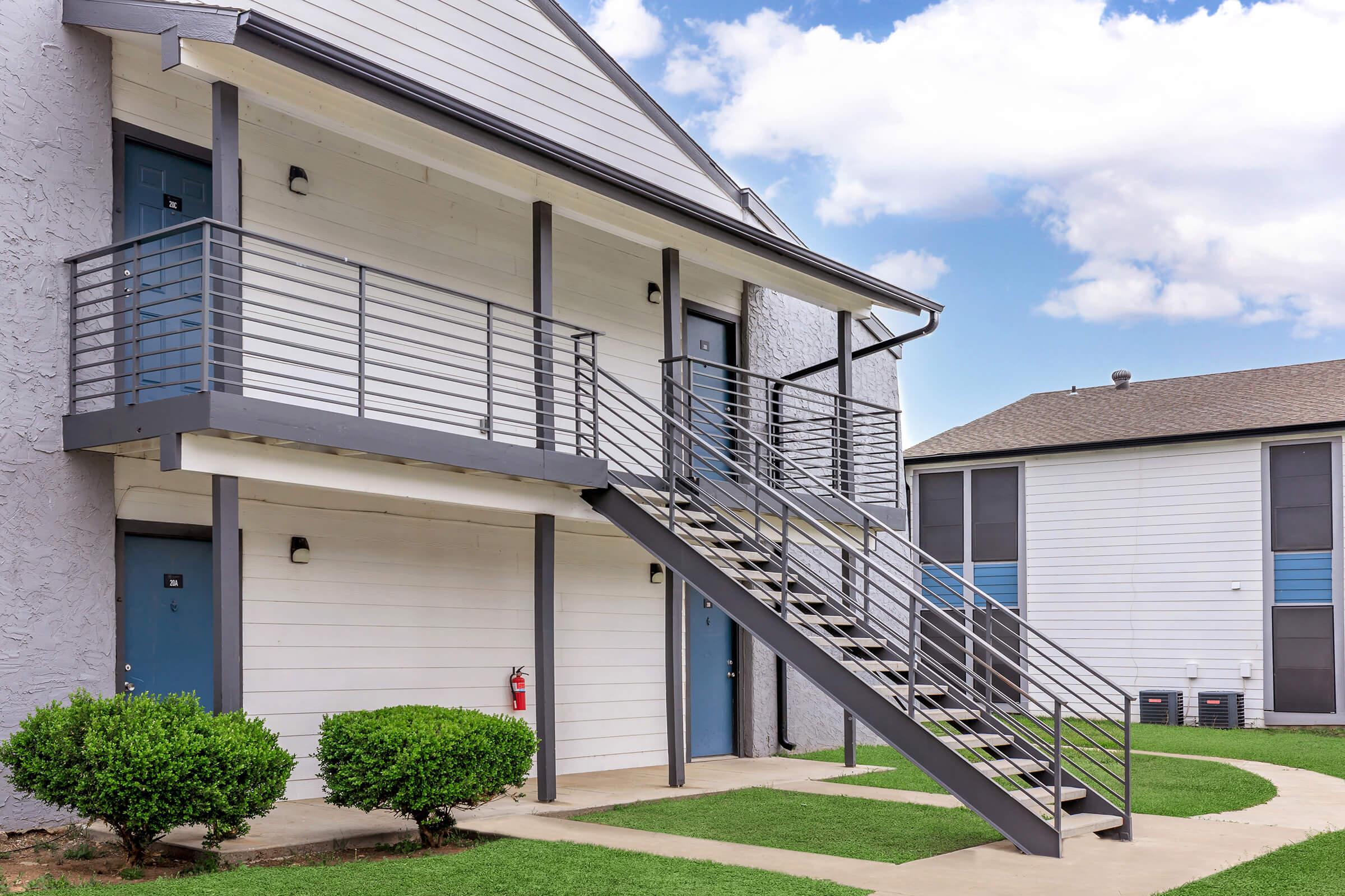 Exterior view of an apartment building featuring a staircase with a metal railing leading to a second floor. The building has light gray siding, blue doors, and small shrubs in front. A fire extinguisher is visible on the ground. Clouds are visible in the sky, indicating a clear day.