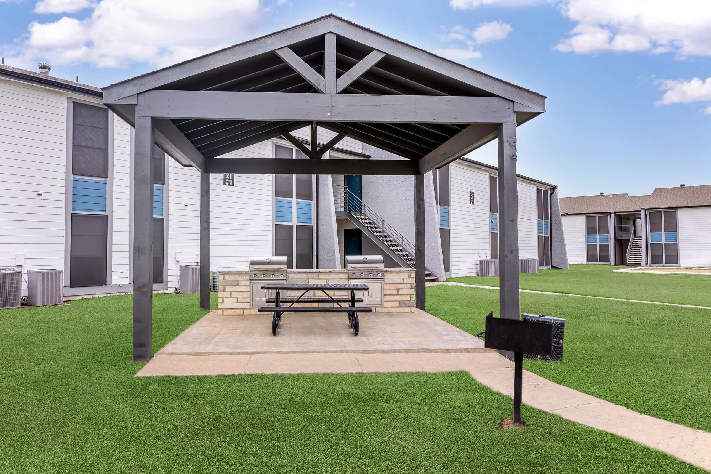 A covered outdoor picnic area with a stone table and benches, surrounded by green grass and near residential buildings. The structures feature modern architecture and large windows, contributing to a welcoming community space.