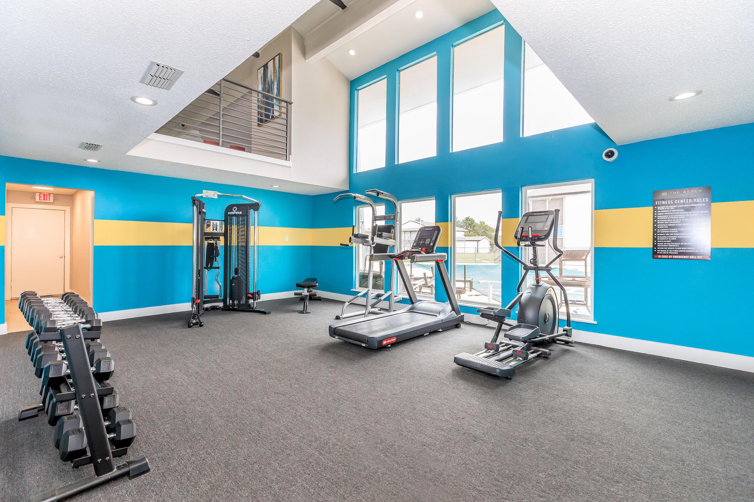 A modern gym interior featuring cardio equipment like treadmills and an elliptical, along with a weightlifting area that has a rack of dumbbells. The walls are painted in blue and yellow, and there are large windows allowing natural light to fill the space.