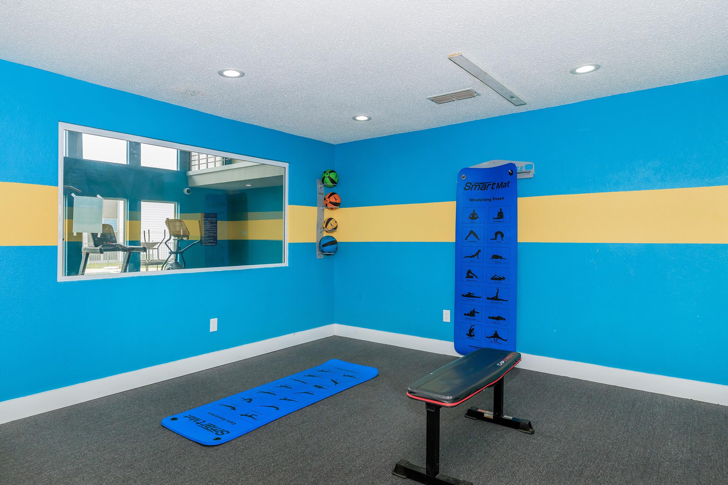 A bright, modern workout space featuring a blue wall with yellow stripes, a blue exercise mat on the floor, a black workout bench, and a large mirror. Wall-mounted fitness equipment is visible, suggesting it's a dedicated area for exercise and training activities.