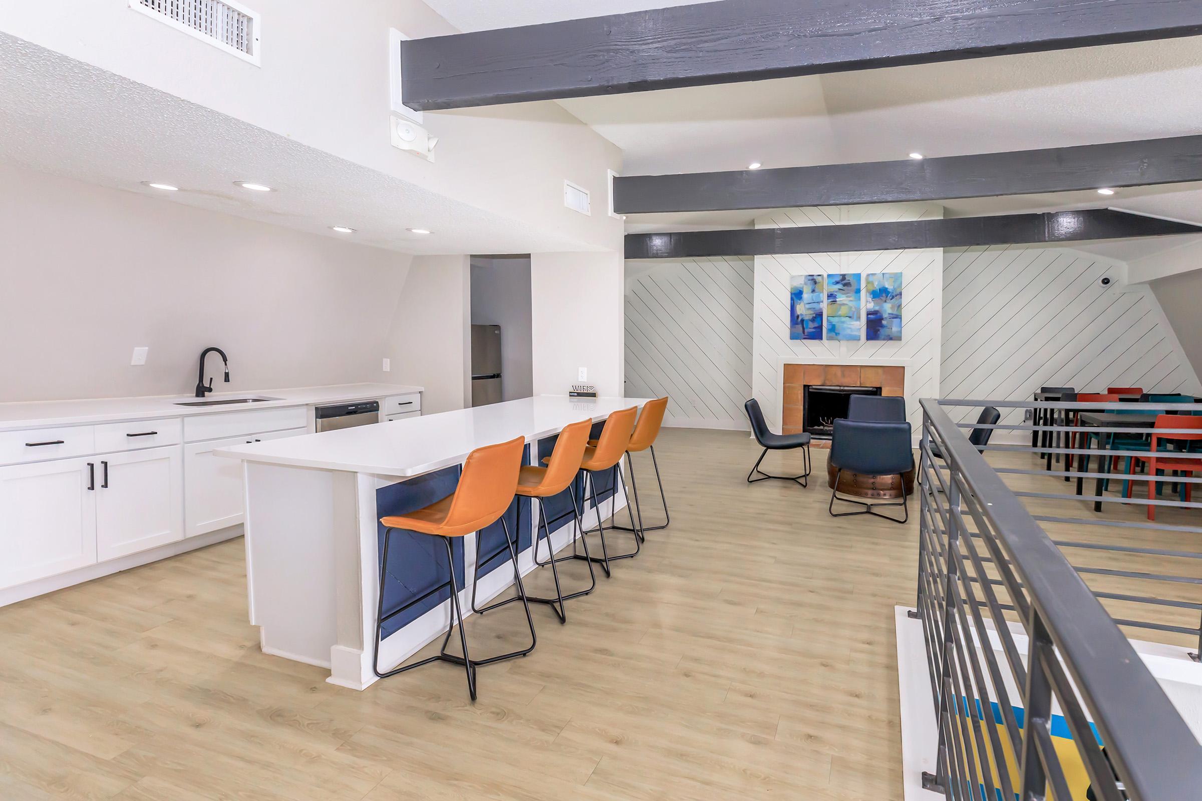 Spacious modern kitchen and living area featuring a large island with bar stools, sleek cabinetry, and a cozy seating area. Natural light streams in from large windows, highlighting the stylish decor, including a fireplace and wall art. Open layout with wooden beams adds to the contemporary vibe.