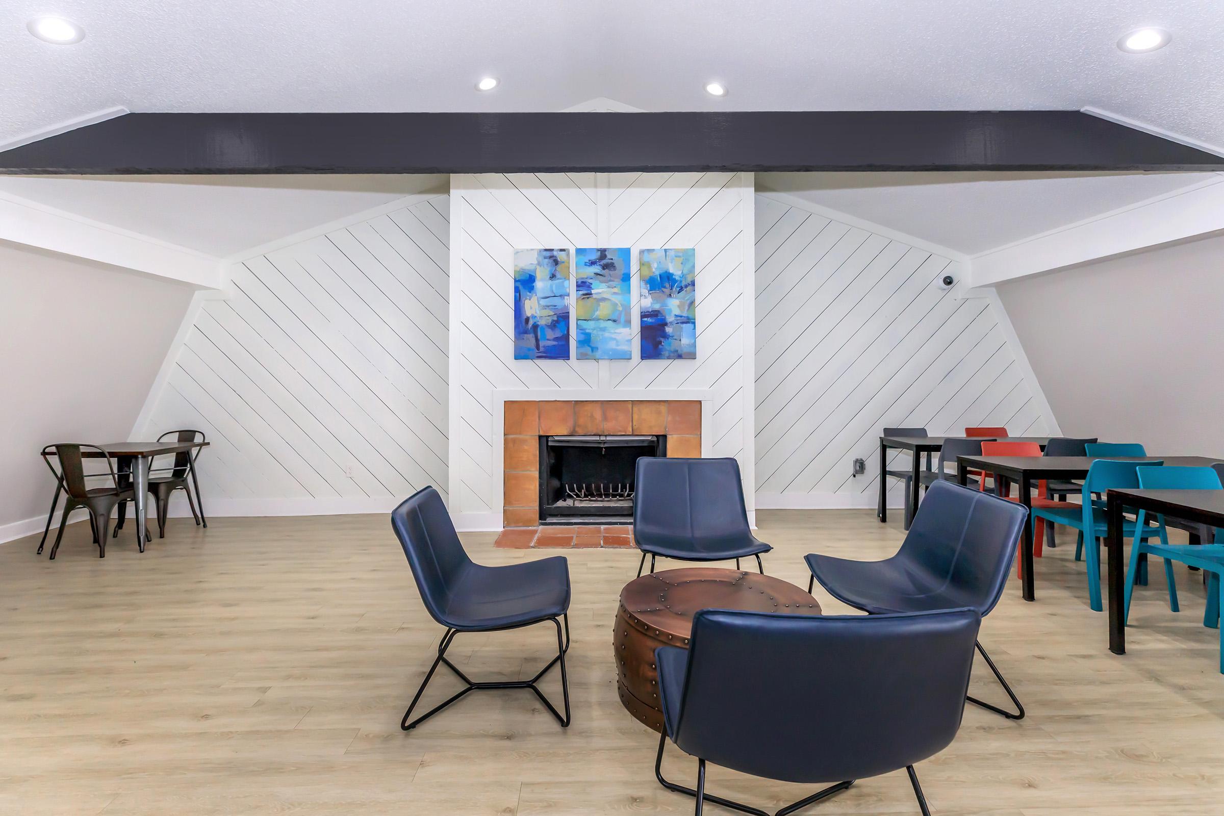 A modern lounge area featuring a stylish fireplace with a brick surround, surrounded by four blue chairs and a circular coffee table. Colorful dining tables in red, blue, and black are also visible, along with artistic blue wall decorations, creating a cozy and inviting atmosphere.