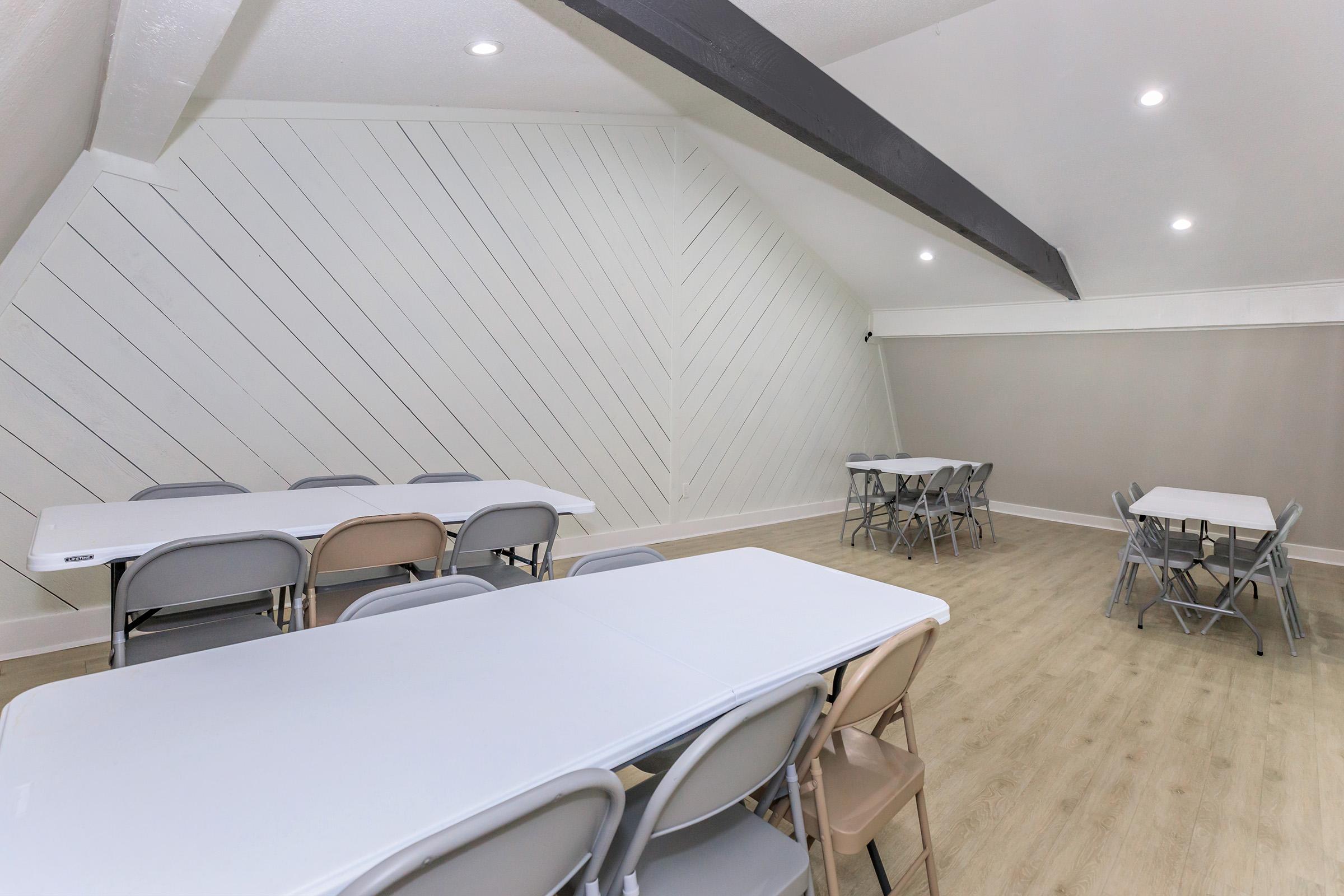 A spacious, well-lit room with light wood flooring. There are several folding tables arranged, with some covered in white tablecloths, and chairs in beige and gray. The walls feature a unique diagonal wood panel design, creating a modern and inviting atmosphere. Ideal for gatherings or meetings.