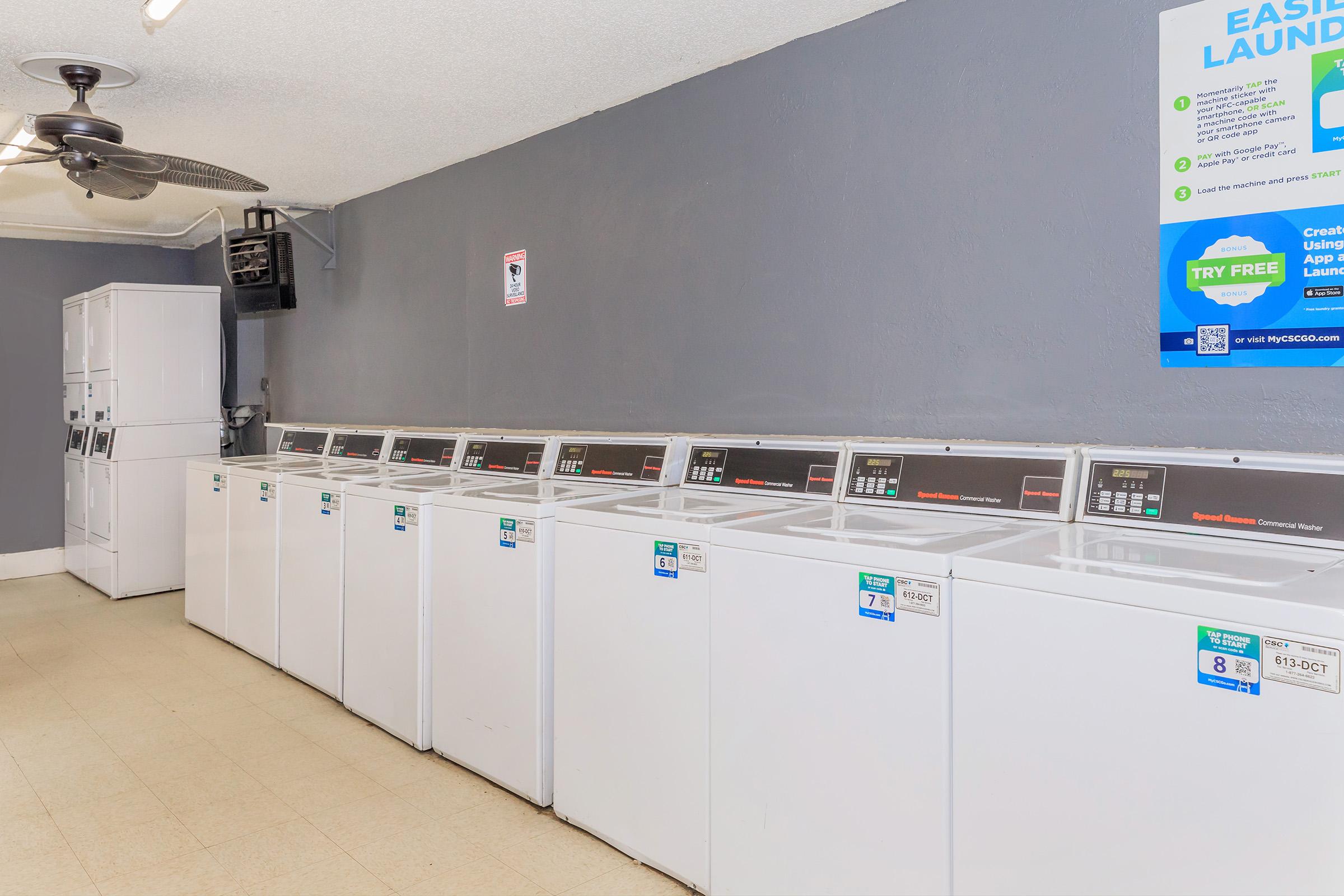 A tidy laundry room featuring several white washing machines lined up against a gray wall. A ceiling fan is visible, and there is a notice board with information, including a sign for an easy laundry service. The floor is light-colored, contributing to a clean and organized appearance.