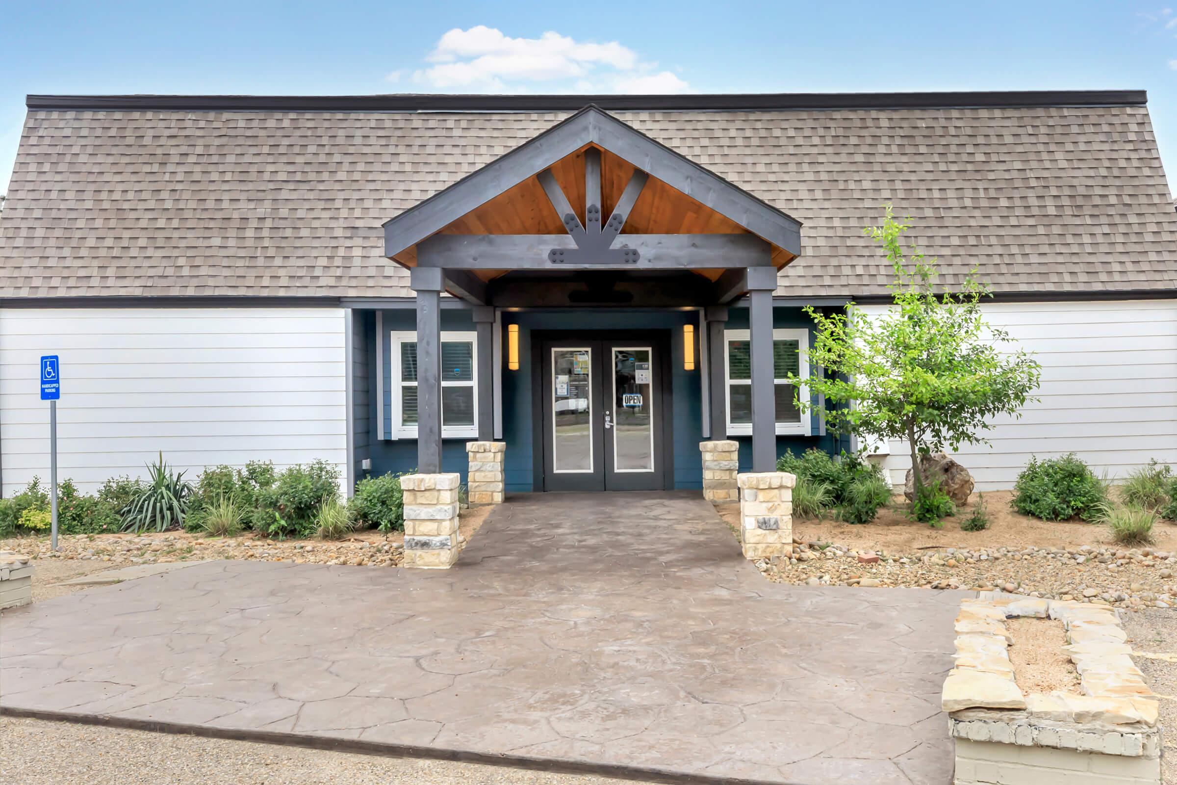A modern building entrance featuring a peaked wooden roof, dark trim, and large glass doors. There are stone accents on either side of the entrance and well-maintained landscaping with shrubs and small trees. A sign indicating accessible parking is visible to the left.