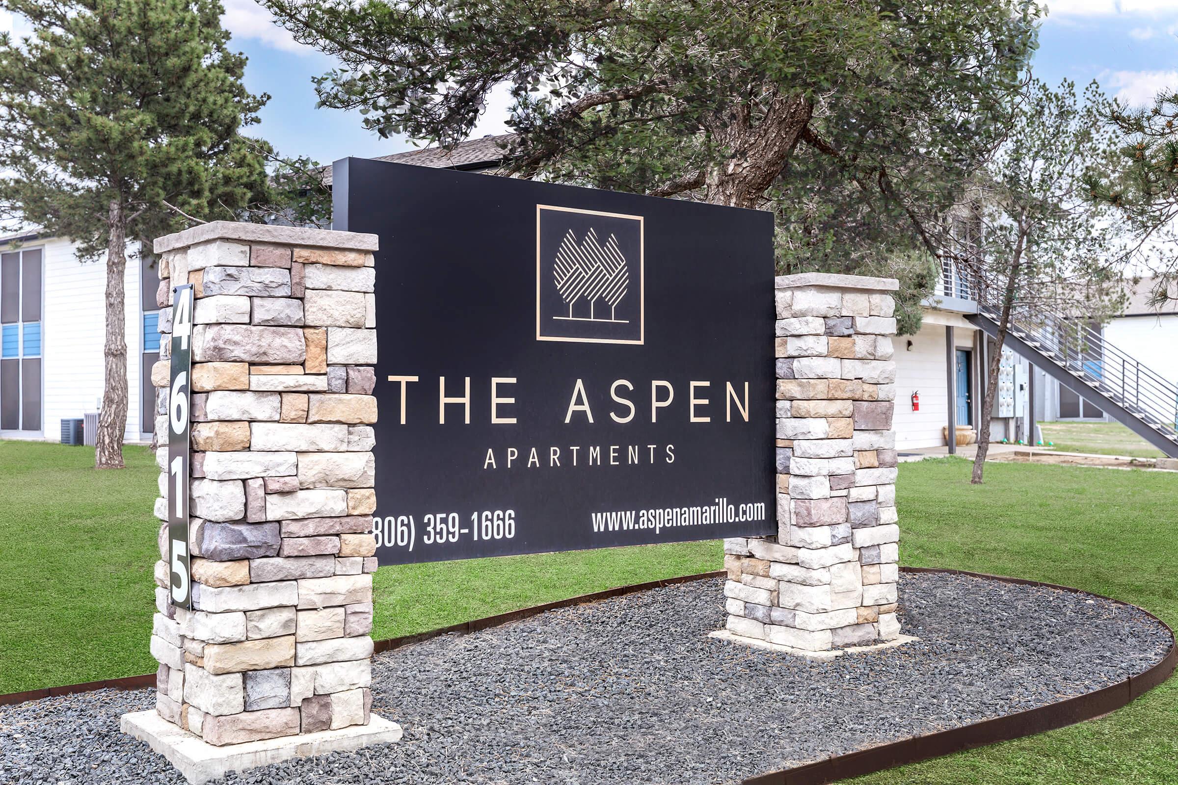 Sign for The Aspen Apartments featuring a modern design with stone pillars and green lawn. The sign displays the apartment name, contact number, and website, showcasing a welcoming residential environment.