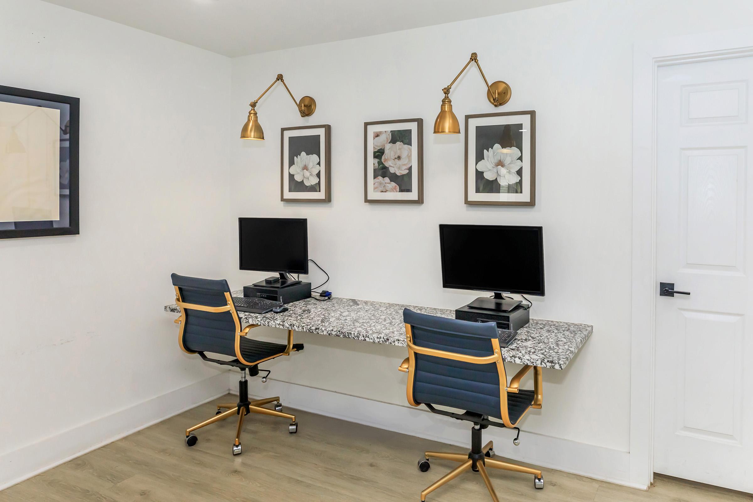Two modern desks with computers are positioned against a white wall. Each desk has a sleek granite surface and gold accents. Above the desks, there are two framed floral prints and stylish wall sconces. The room features light wood flooring and a minimalist design.
