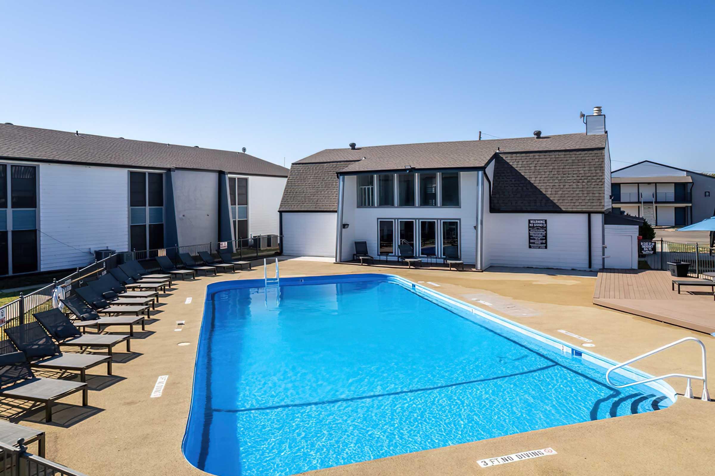 A clear blue swimming pool surrounded by lounge chairs and a deck, with a two-story building in the background. The sky is bright and sunny, highlighting the inviting atmosphere of the area.