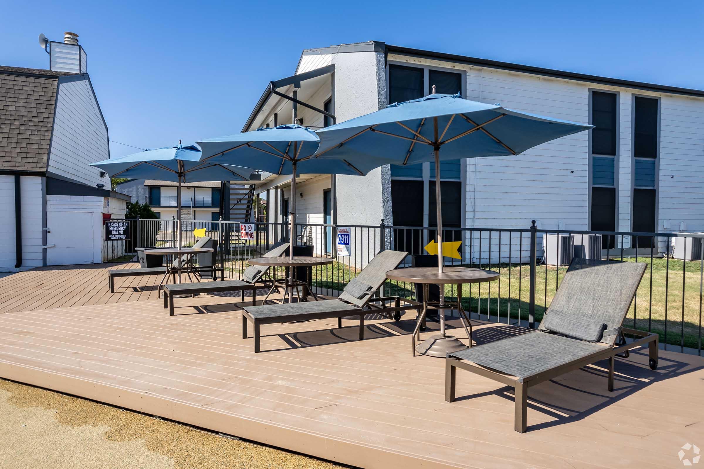 Outdoor seating area with several lounge chairs and tables shaded by blue umbrellas. The setting features a wooden deck, surrounded by grass, and a multi-story building in the background under a clear blue sky.