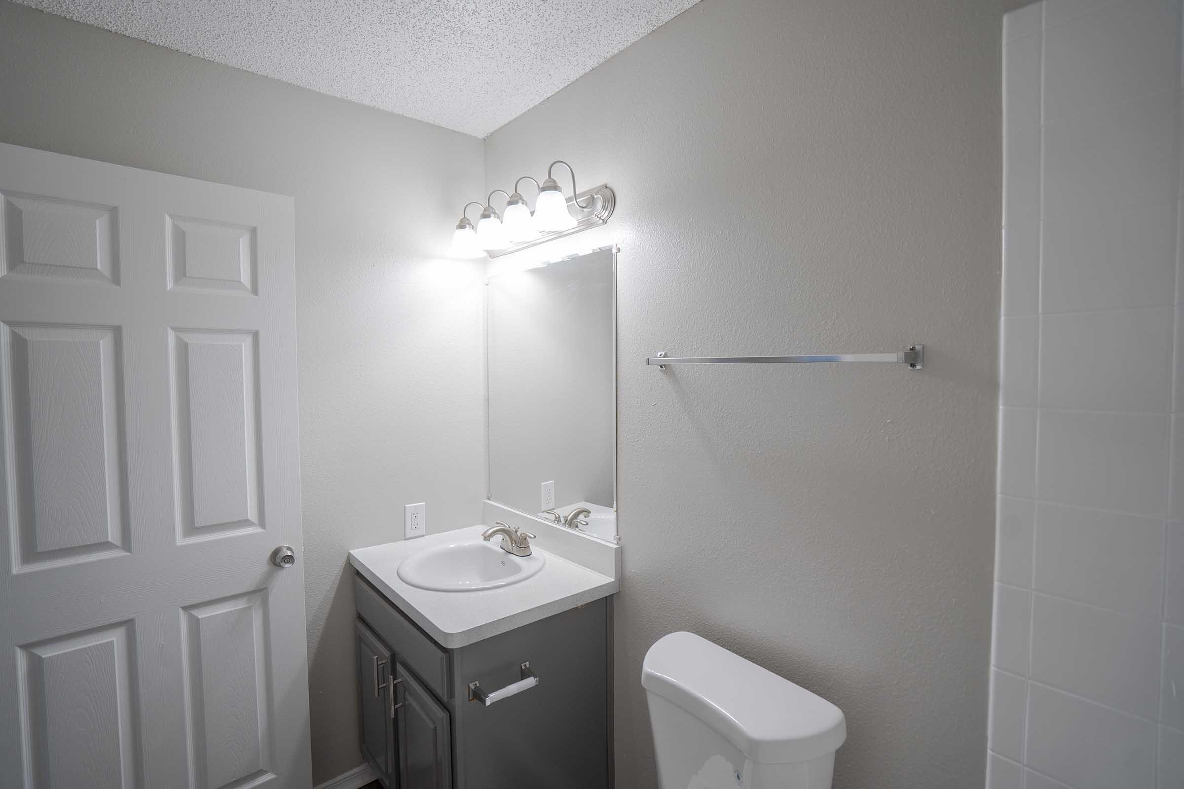 A well-lit bathroom featuring a white sink with a silver faucet, a large mirror above it, a towel rack, and a toilet. The walls are painted a light gray, and there is a white shower area visible in the corner. A closed white door is also present in the image, adding to the overall clean and modern aesthetic.
