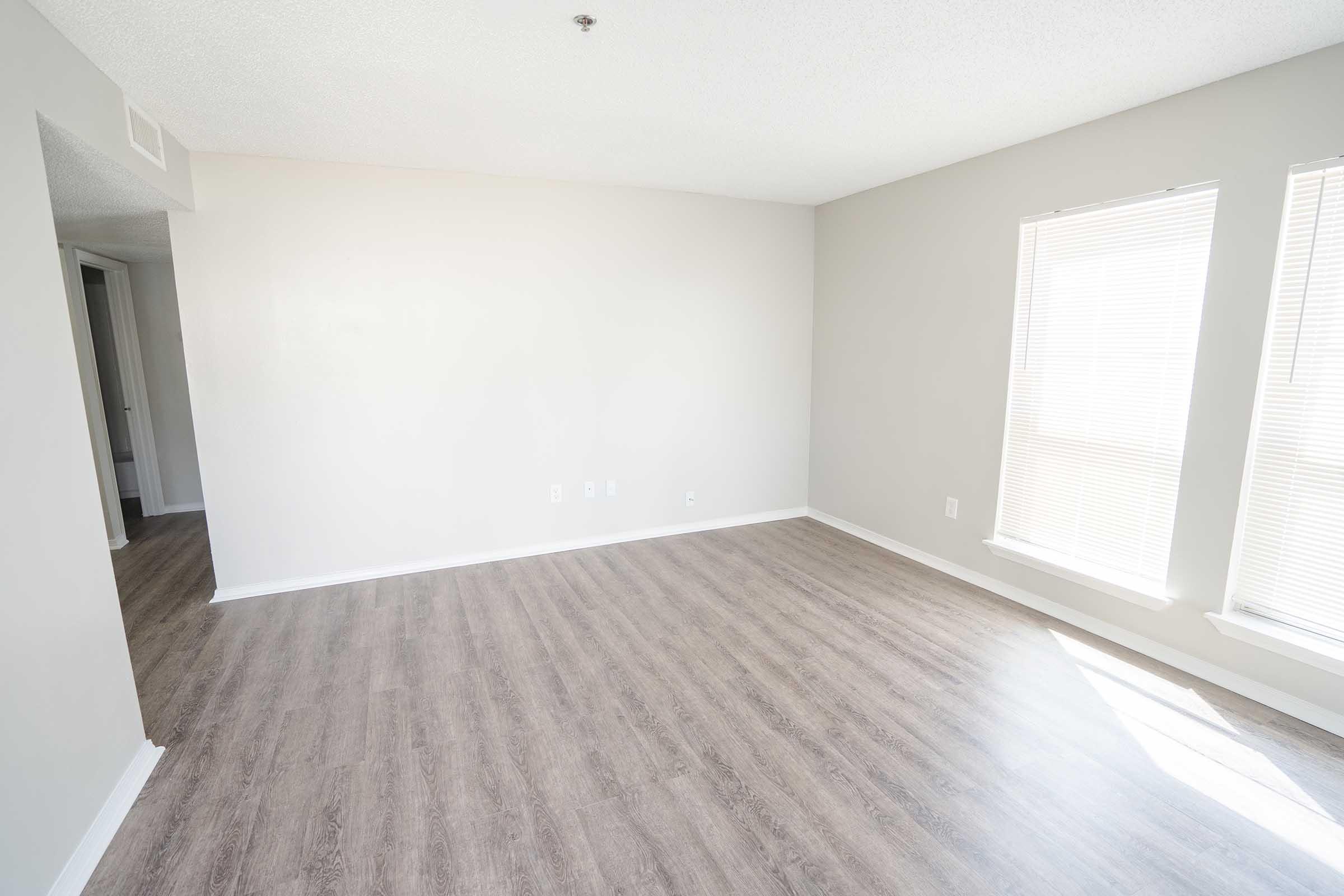 A spacious, empty room with light-colored walls and laminate flooring. Two windows with blinds allow natural light to enter. The room features a door leading to another area, creating an open and inviting atmosphere.