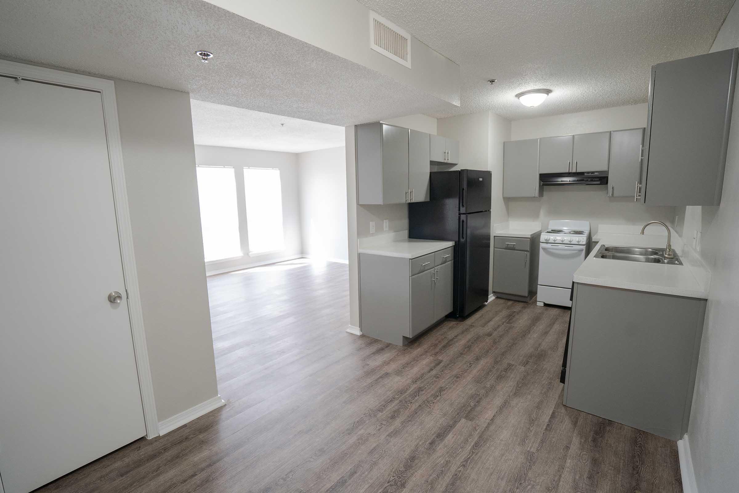 A modern kitchen featuring gray cabinets, a black refrigerator, and a white stove. The open layout connects to a bright living area with large windows. The flooring is a light wood laminate, and the walls are painted in a neutral color.