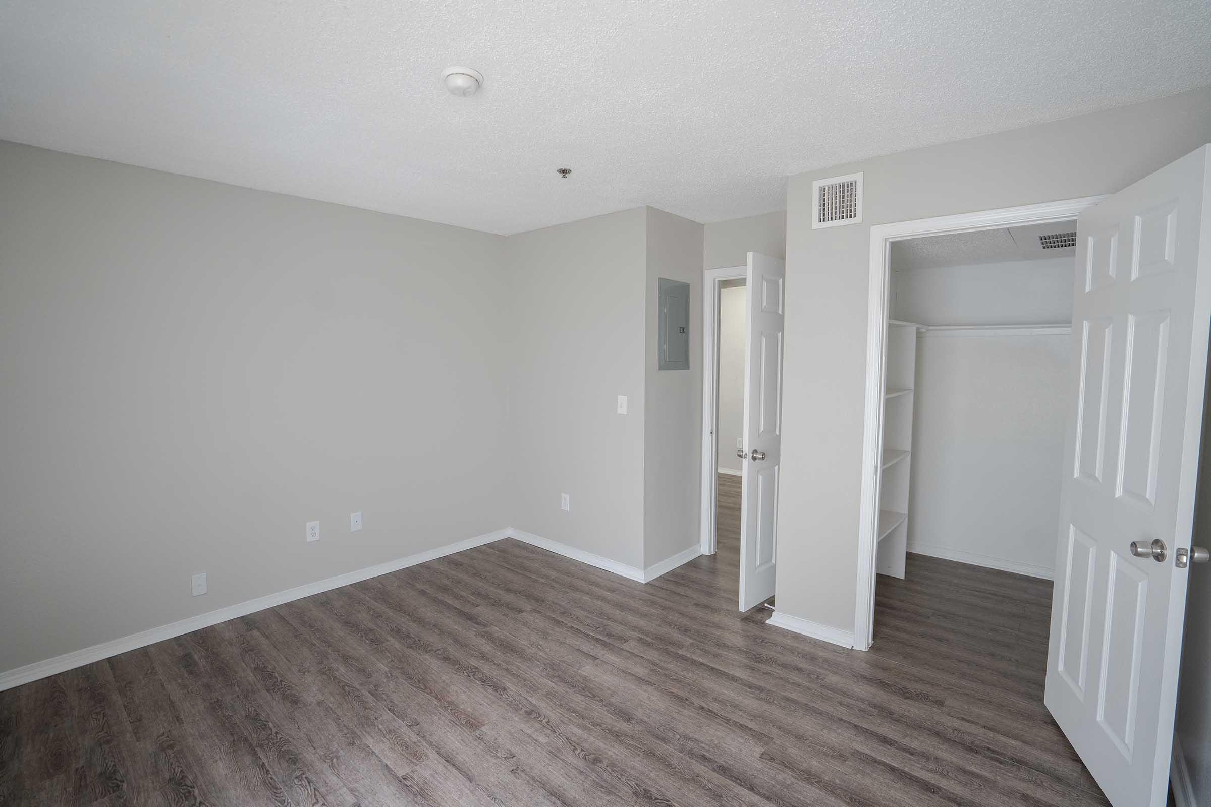A spacious, empty room with light gray walls and a textured ceiling. It features laminate flooring, two doors leading to adjacent rooms, and a closet with shelving. Natural light reflects off the walls, creating a bright atmosphere.