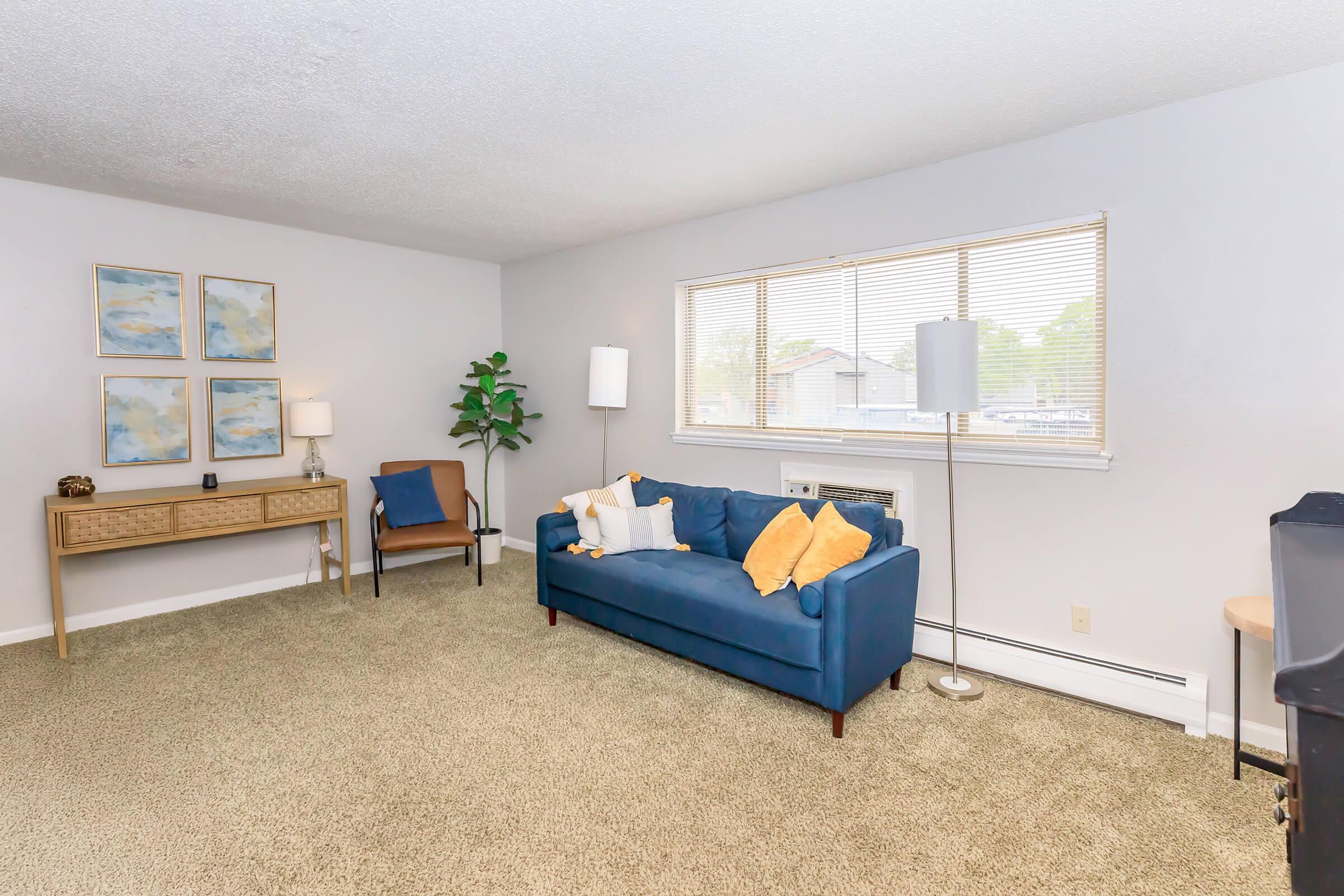 A cozy living room featuring a blue sofa with yellow pillows, a wooden console table, an armchair, and a potted plant. There's a window letting in natural light and two lamps providing additional illumination. The walls are light gray, and the floor is covered with a plush carpet.