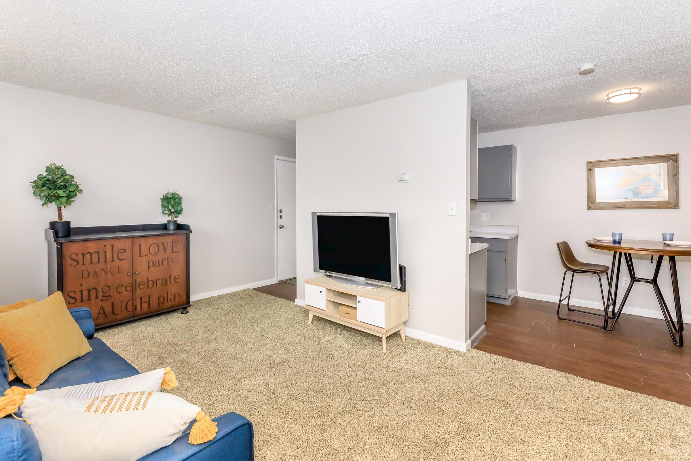 Cozy living area featuring a blue sofa with yellow pillows, a wall-mounted TV on a light wood stand, and a decorative bar with inspirational text. The adjacent dining area includes a wooden table with chairs and a framed picture on the wall. Warm carpet and soft lighting create a welcoming atmosphere.