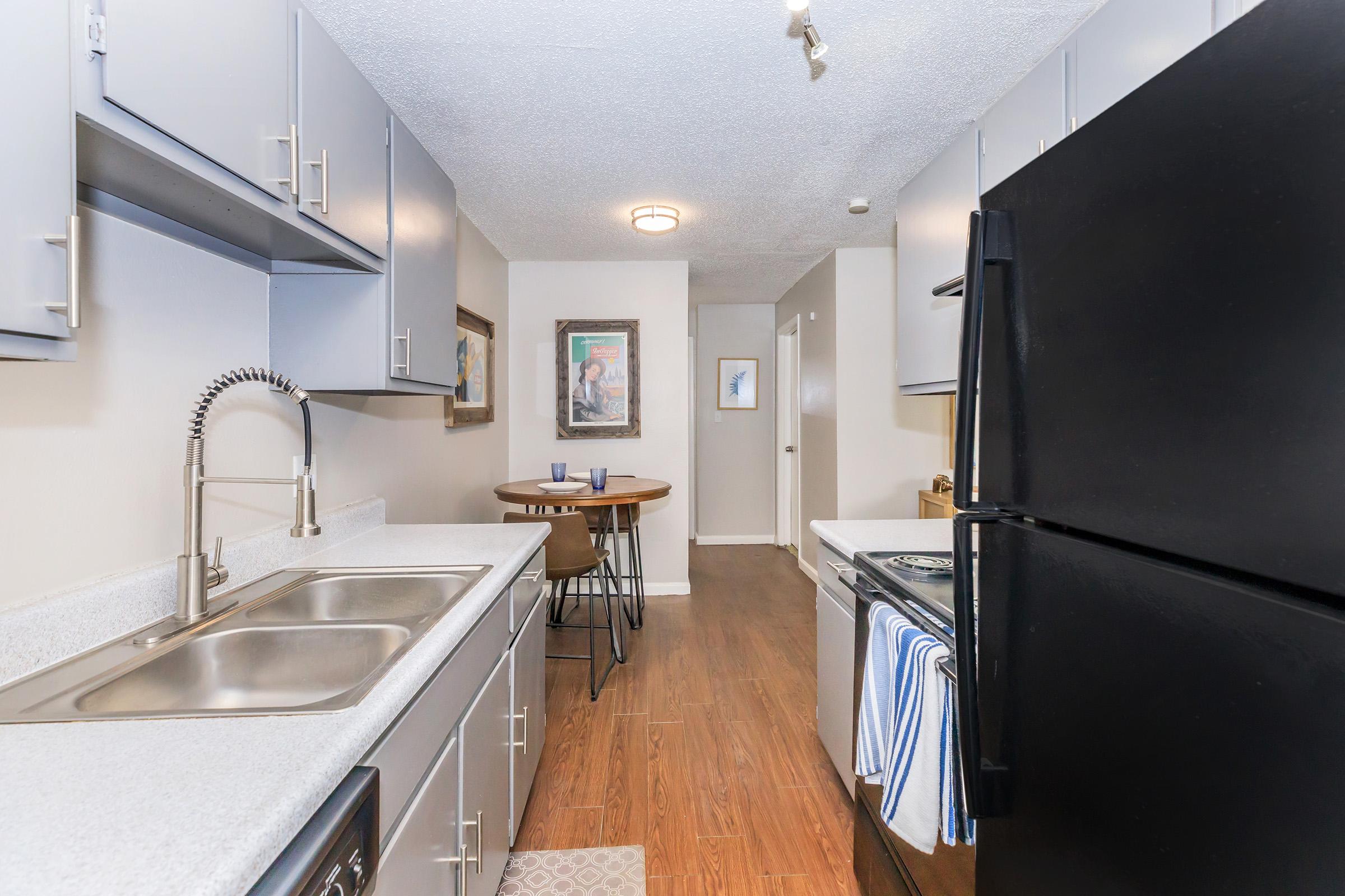 A modern kitchen with gray cabinets, a double sink, and a black refrigerator. The countertop is light-colored, and there’s a small round dining table with two chairs in the corner. The flooring is wooden, and there's a light fixture on the ceiling. Wall art is visible in the background.