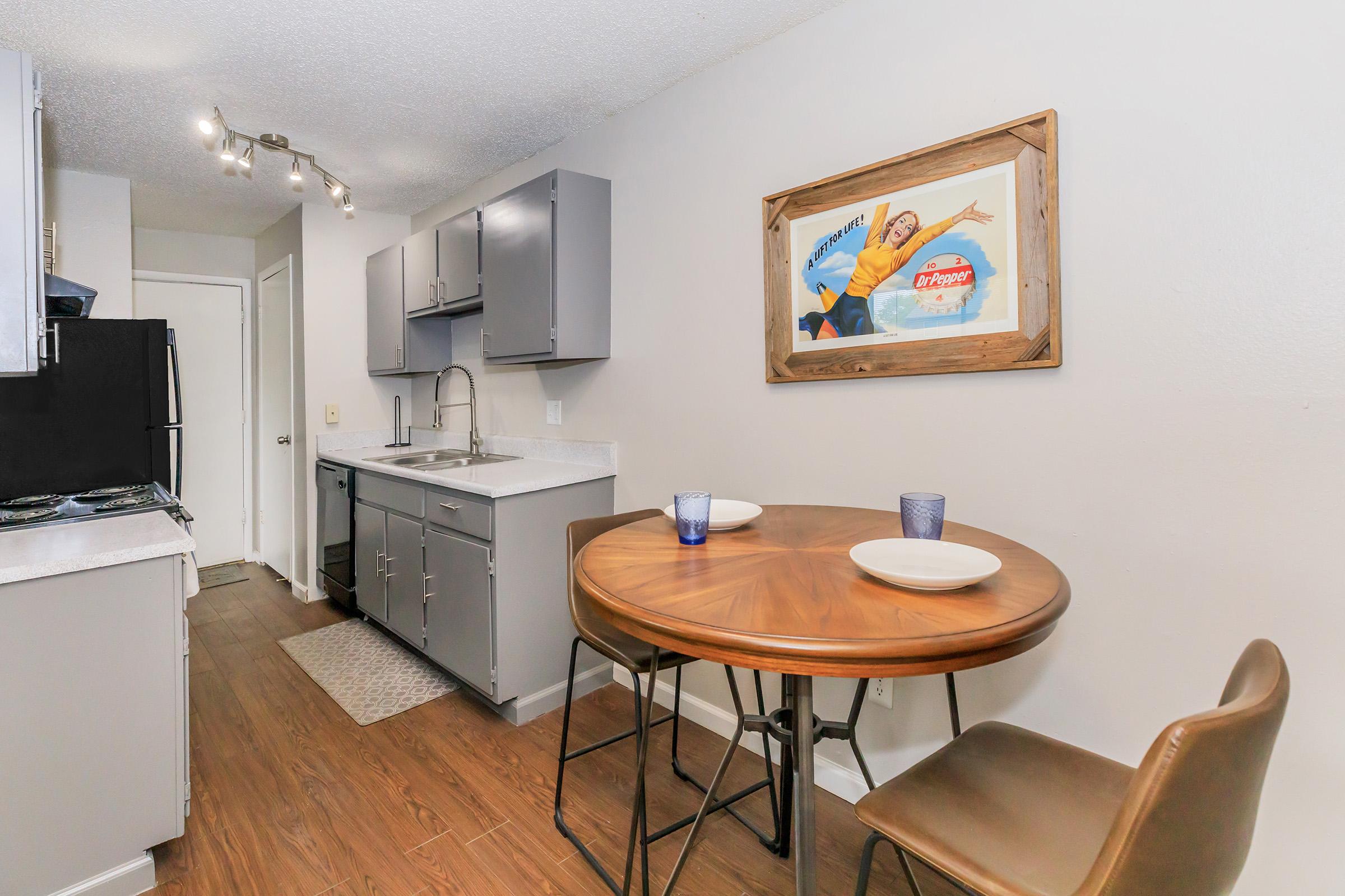 A modern kitchen featuring grey cabinets, a sink, and countertop. There is a round wooden table with two blue glasses and plates, surrounded by stylish chairs. A vintage-style framed artwork is on the wall. The space has warm tones and a clean, contemporary design.