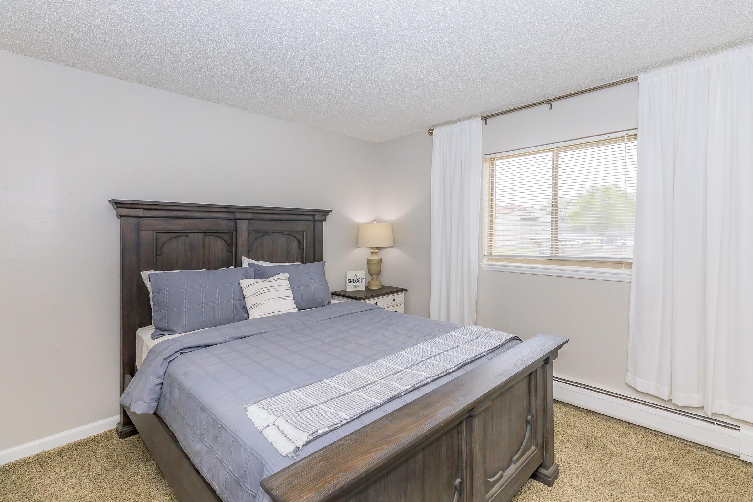 A cozy bedroom featuring a dark wooden bed with gray bedding, a bedside table with a lamp, and a large window dressed with white curtains. The walls are painted light gray, and the floor is carpeted in a neutral tone, creating a warm and inviting atmosphere.