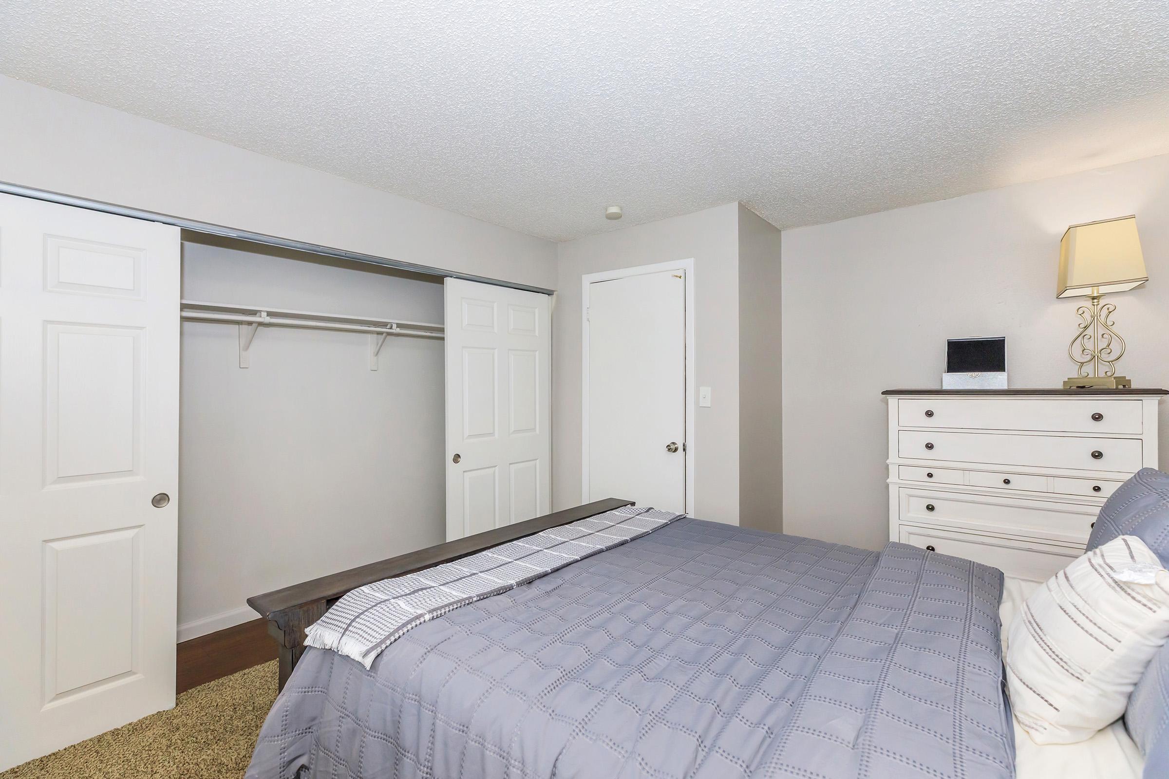 A cozy bedroom featuring a neatly made bed with gray bedding, a white dresser, and a lamp. There are two closet doors on the left, one of which is open, revealing a hanging rod. The walls are painted a light color, and the carpeted floor adds warmth to the space.