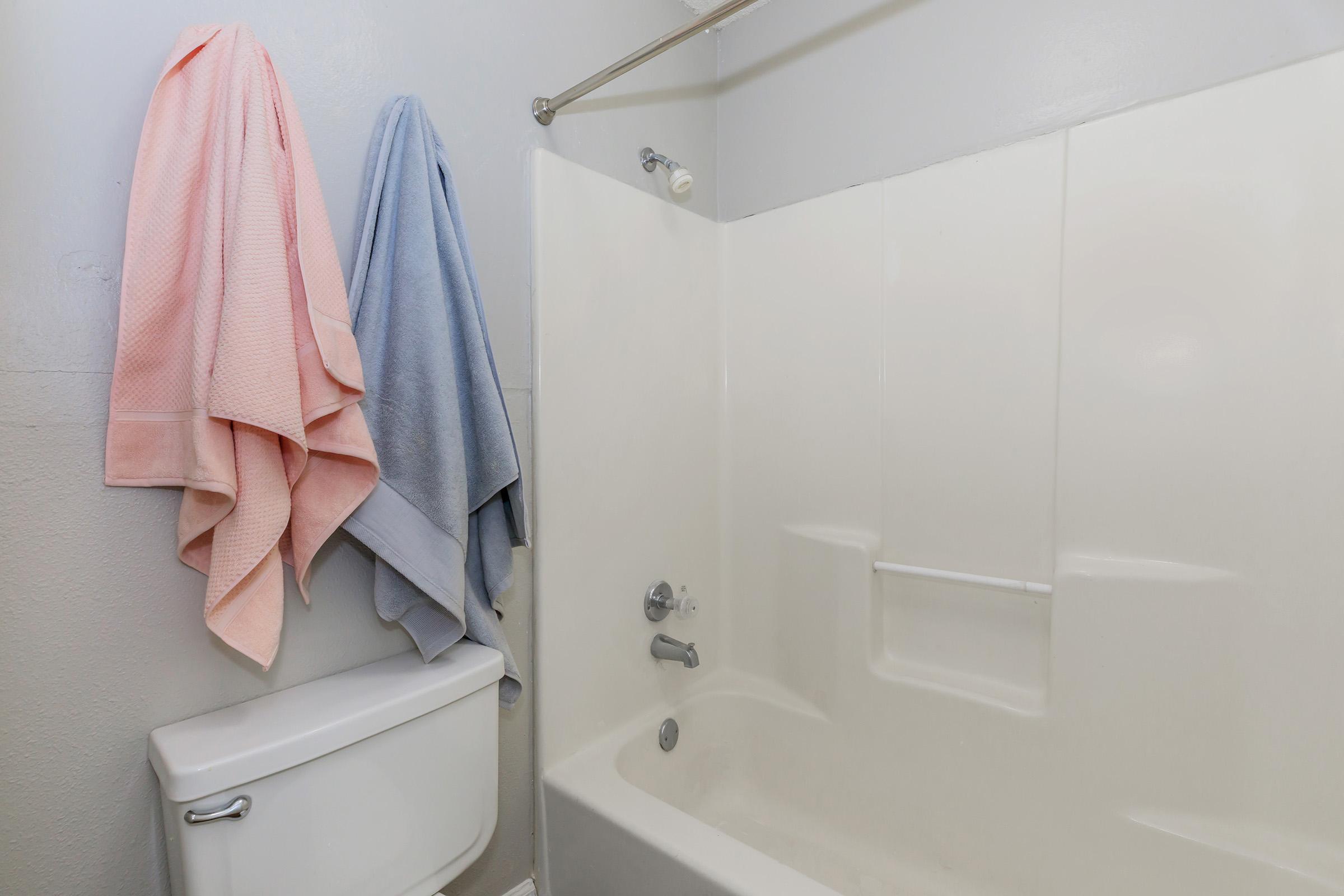 A bathroom featuring a white bathtub with a showerhead, a white toilet nearby, and two hanging towels—one pink and one light blue—draped on a metal rod. The walls are a light color, creating a clean and simple atmosphere.