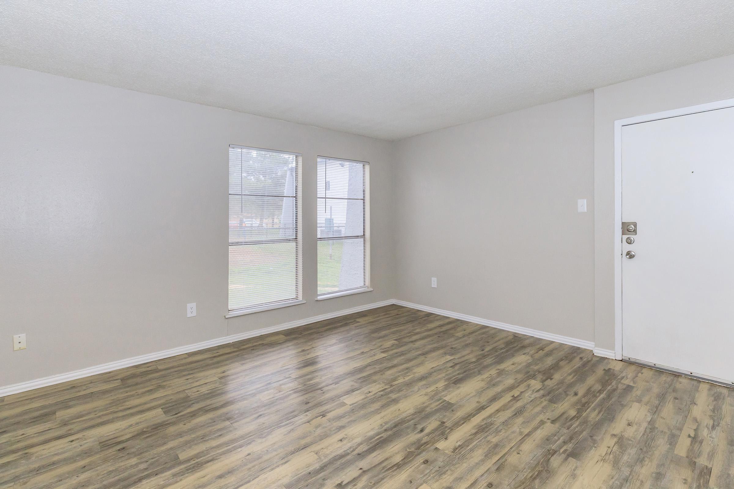 Interior view of a spacious, empty room with light gray walls and a textured ceiling. It features large windows allowing natural light, a smooth floor in a wood-like finish, and a white door leading outside. The overall atmosphere is bright and inviting, ideal for decorating or furnishing.