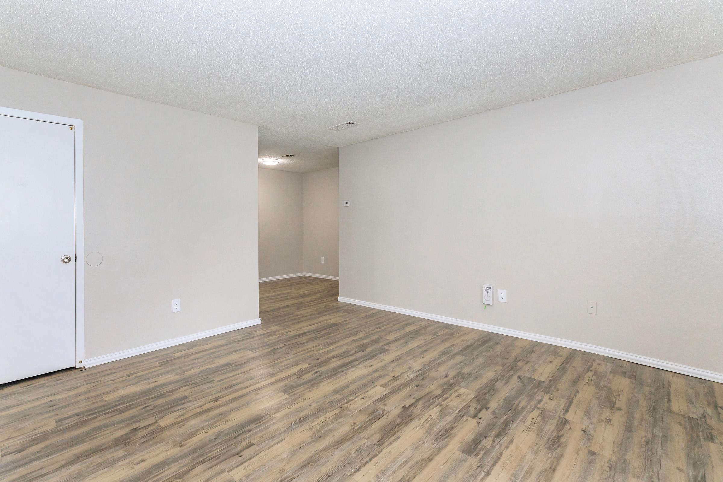 Empty room with light-colored walls and a hardwood floor. A white door is visible on the left, and the room has a neutral, uncluttered appearance. The space is well-lit, and there is a doorway leading to another room in the background.
