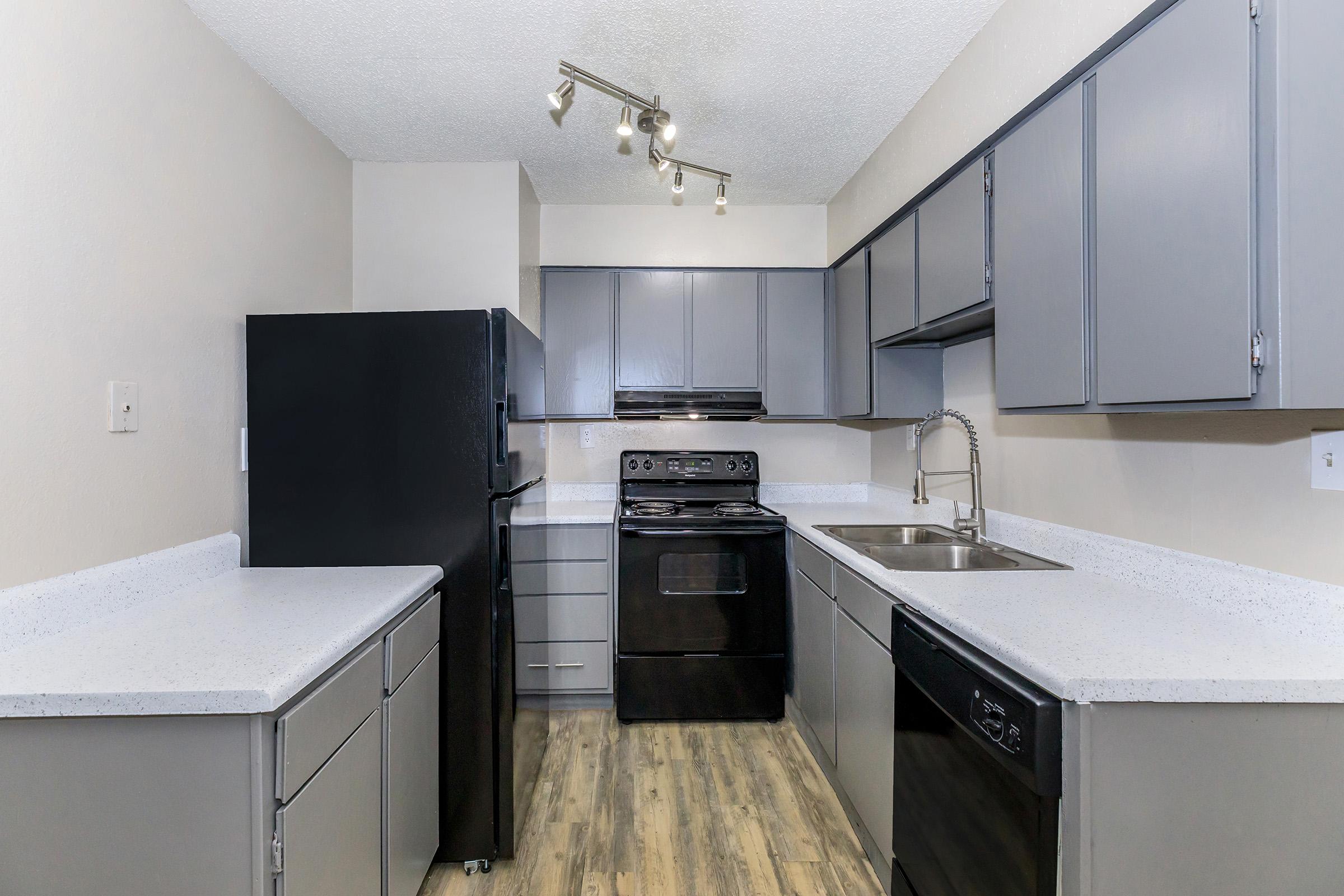 Modern kitchen featuring gray cabinetry, a black refrigerator, and a black oven. The countertops are a light color, and there are stainless steel fixtures. The space has a clean, contemporary design with overhead lighting and a neutral wall color, creating a bright and open atmosphere.