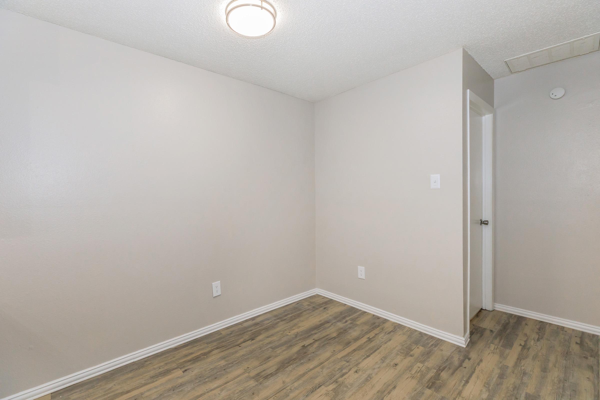 A vacant room with light-colored walls, wooden flooring, a ceiling light fixture, and a door on the right leading to another space. The room has minimal decor and is empty, creating a simple and clean aesthetic.