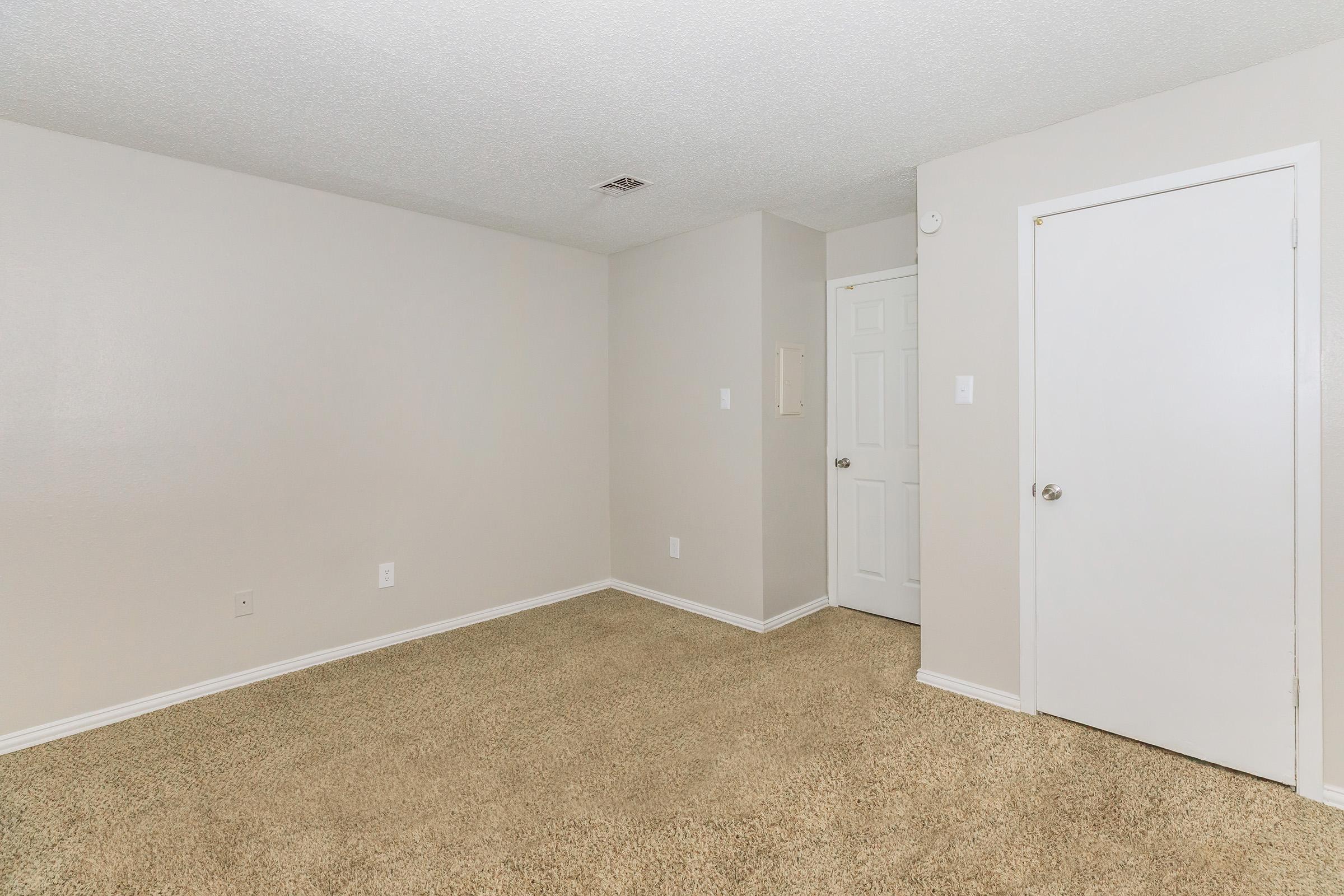 A empty room with light beige carpeting and plain beige walls. There is a closed white door on the right and another doorframe leading to an adjacent space, with no furniture or decorations visible. The ceiling is textured and has a few light fixtures.