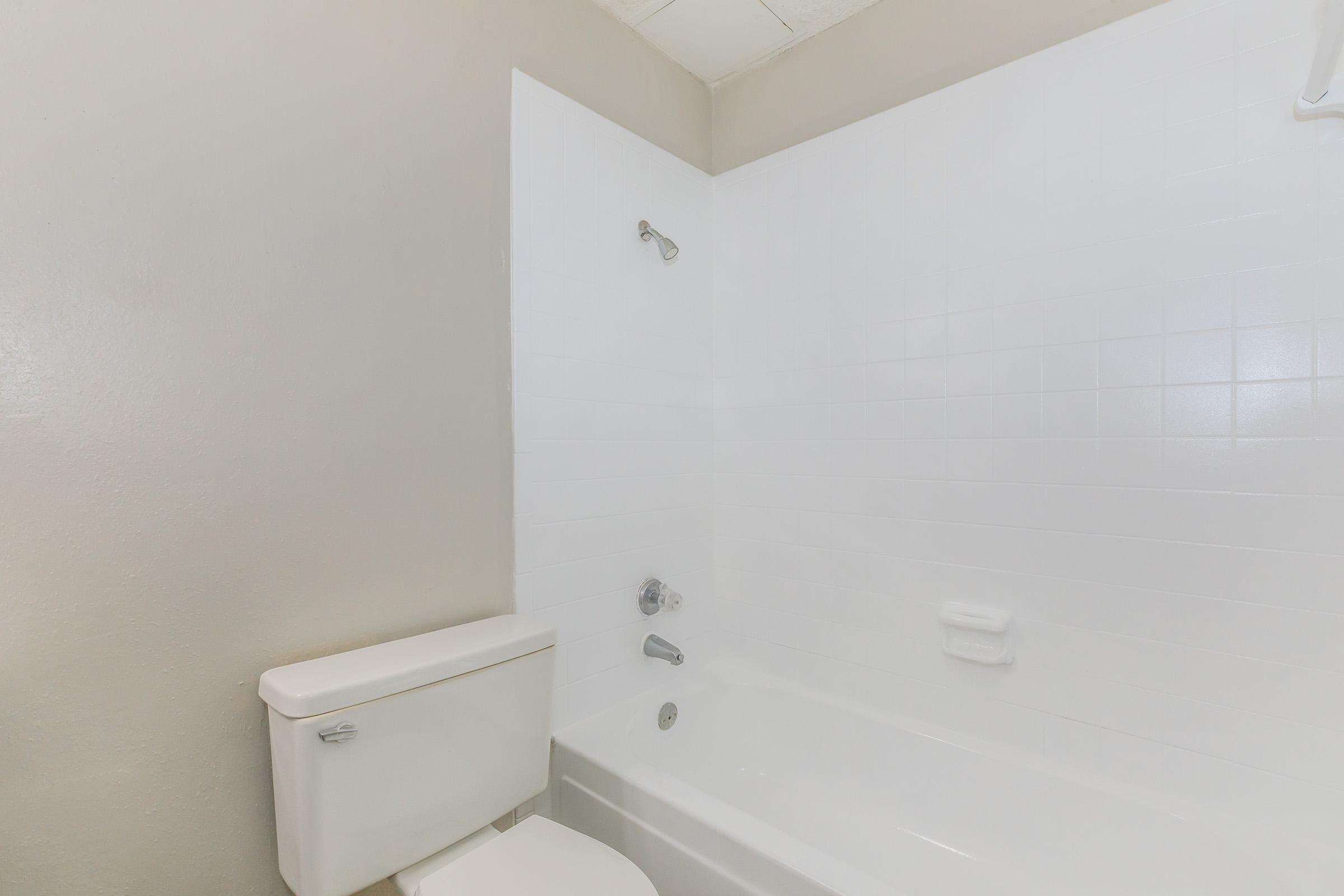 A clean, simple bathroom featuring a white bathtub with a showerhead, a toilet beside it, and light gray walls. The space is well-lit and modern, with basic fixtures.
