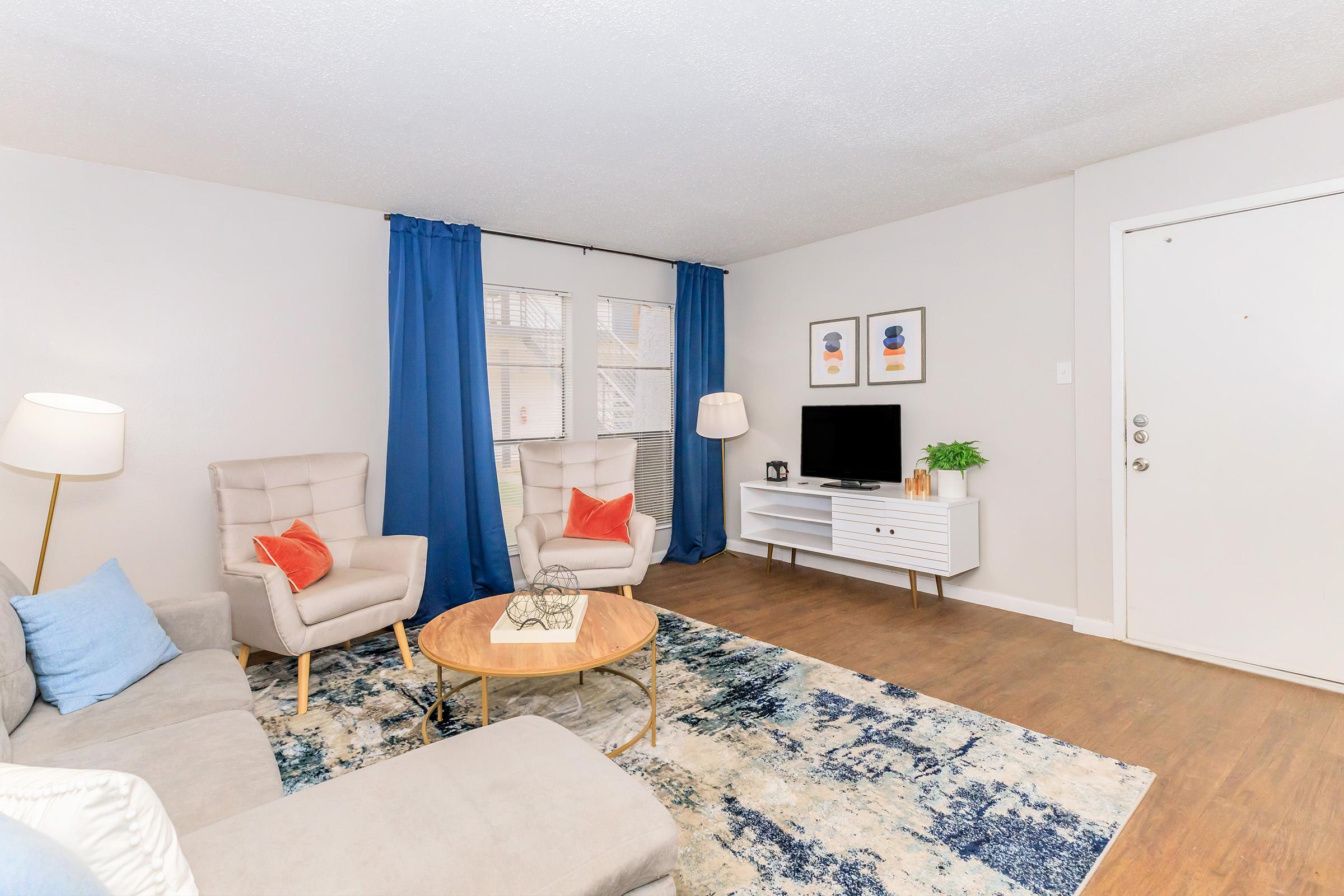 A cozy living room featuring two light gray armchairs, a light gray sectional sofa, a round wooden coffee table, and a white media console with a TV. The windows are dressed with blue curtains, and there’s a decorative rug with blue and white patterns on the floor. A welcoming atmosphere in soft, neutral tones.