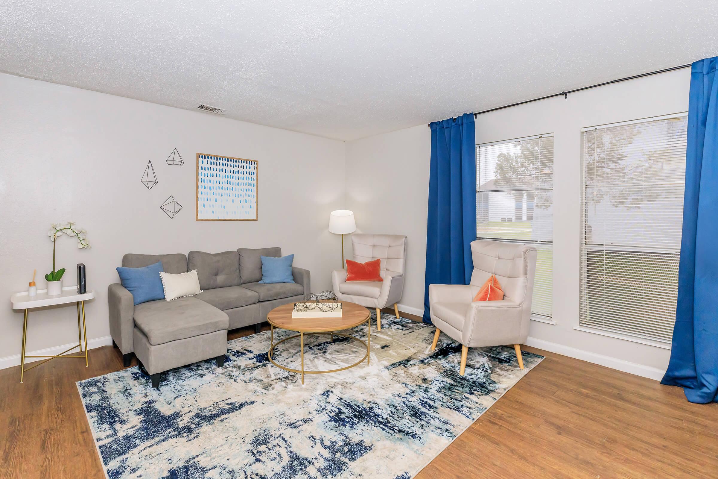 A modern living room featuring a gray sofa with blue and orange accent pillows, two light-colored armchairs with orange cushions, a round wooden coffee table on a patterned blue rug, and a side table with a plant. Large windows with blue curtains allow natural light to fill the space. Decorative wall art complements the decor.