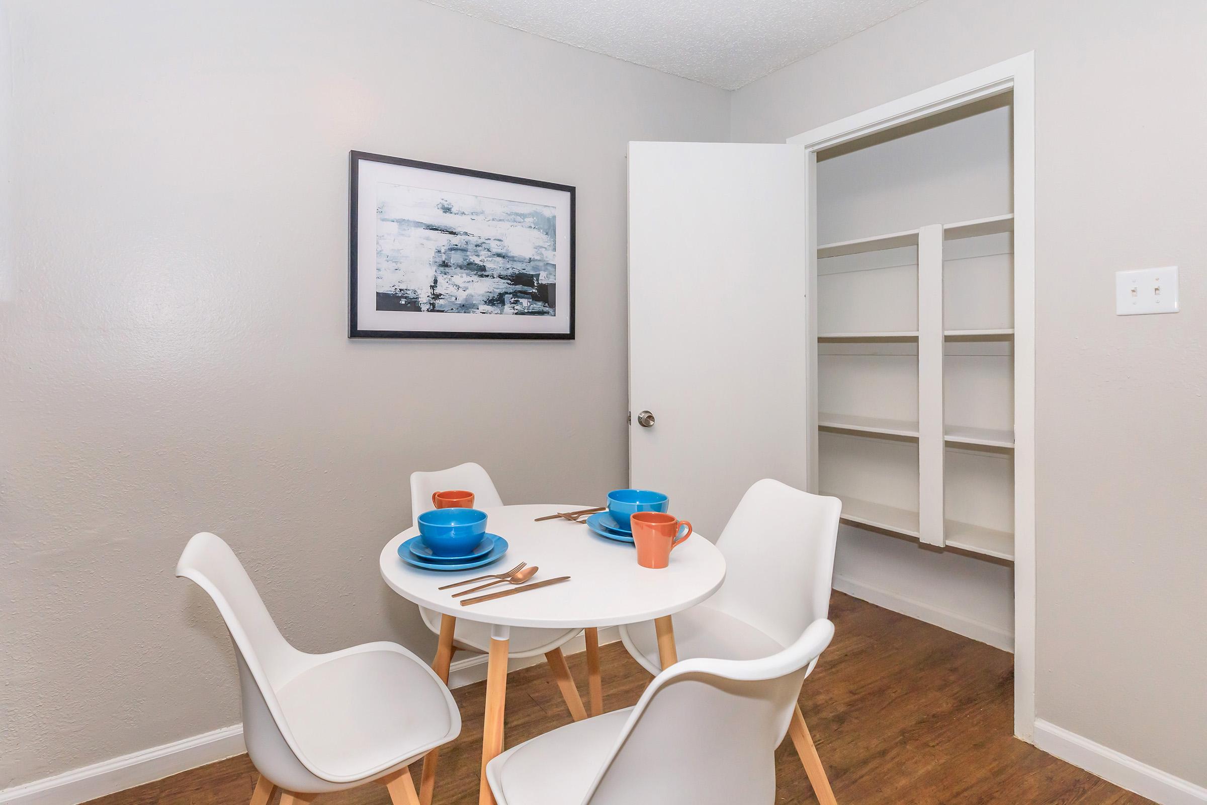 A bright dining area featuring a round white table with four chairs. The table is set with blue and terracotta dishes and cutlery. A framed abstract painting hangs on the wall, and a doorway leads to a simple storage area with open shelves. The flooring is a warm wood finish.