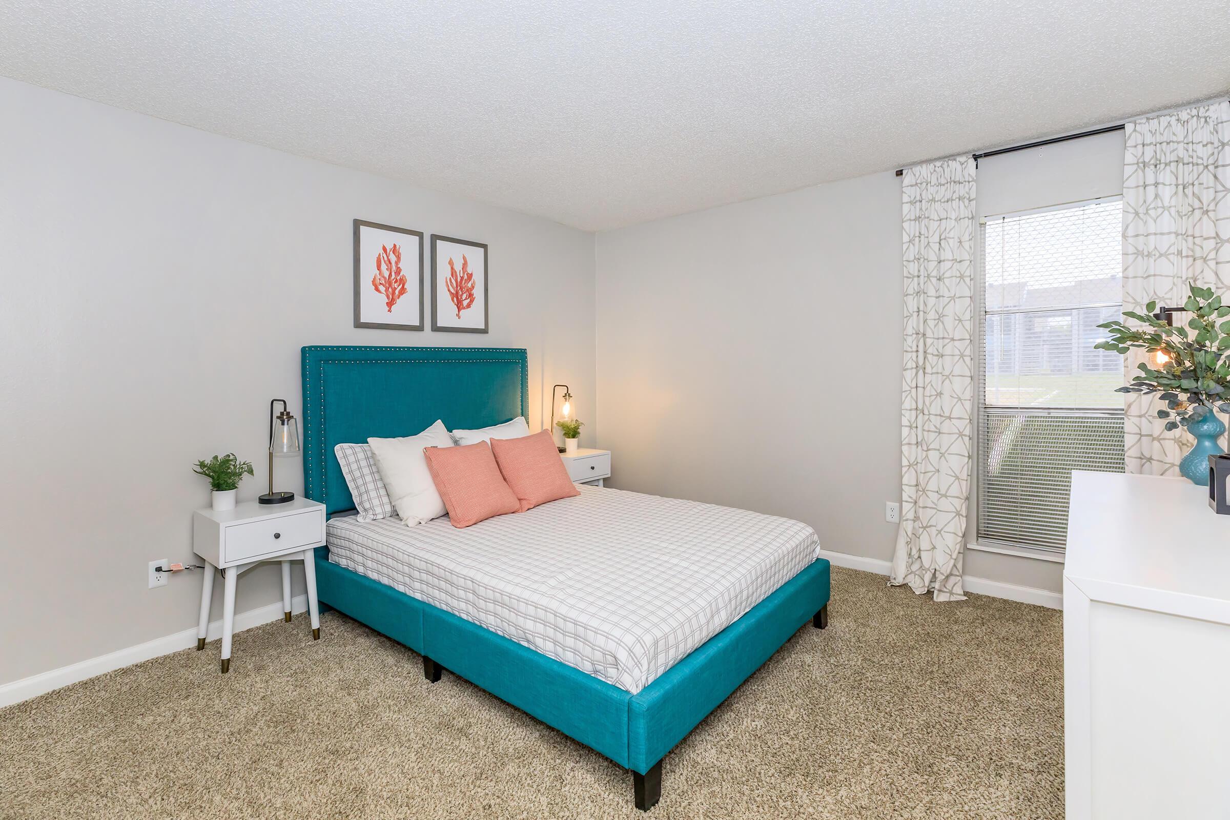 A modern bedroom featuring a teal upholstered bed with white bedding and coral accent pillows. Beside the bed are two white nightstands with lamps. The walls are light gray, adorned with coral-themed artwork. A large window with sheer curtains allows natural light to fill the room, complemented by a decorative plant on a dresser.