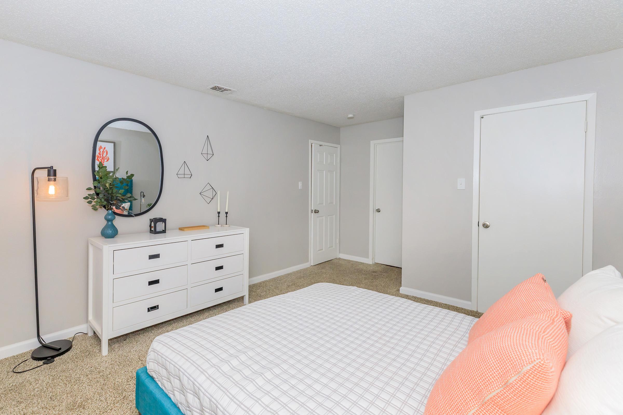 A well-decorated bedroom featuring a white bed with orange throw pillows. There's a white dresser with a mirror above, an accent lamp, and a decorative plant. The walls are light gray and there are two doors visible, enhancing the modern and cozy atmosphere of the room.