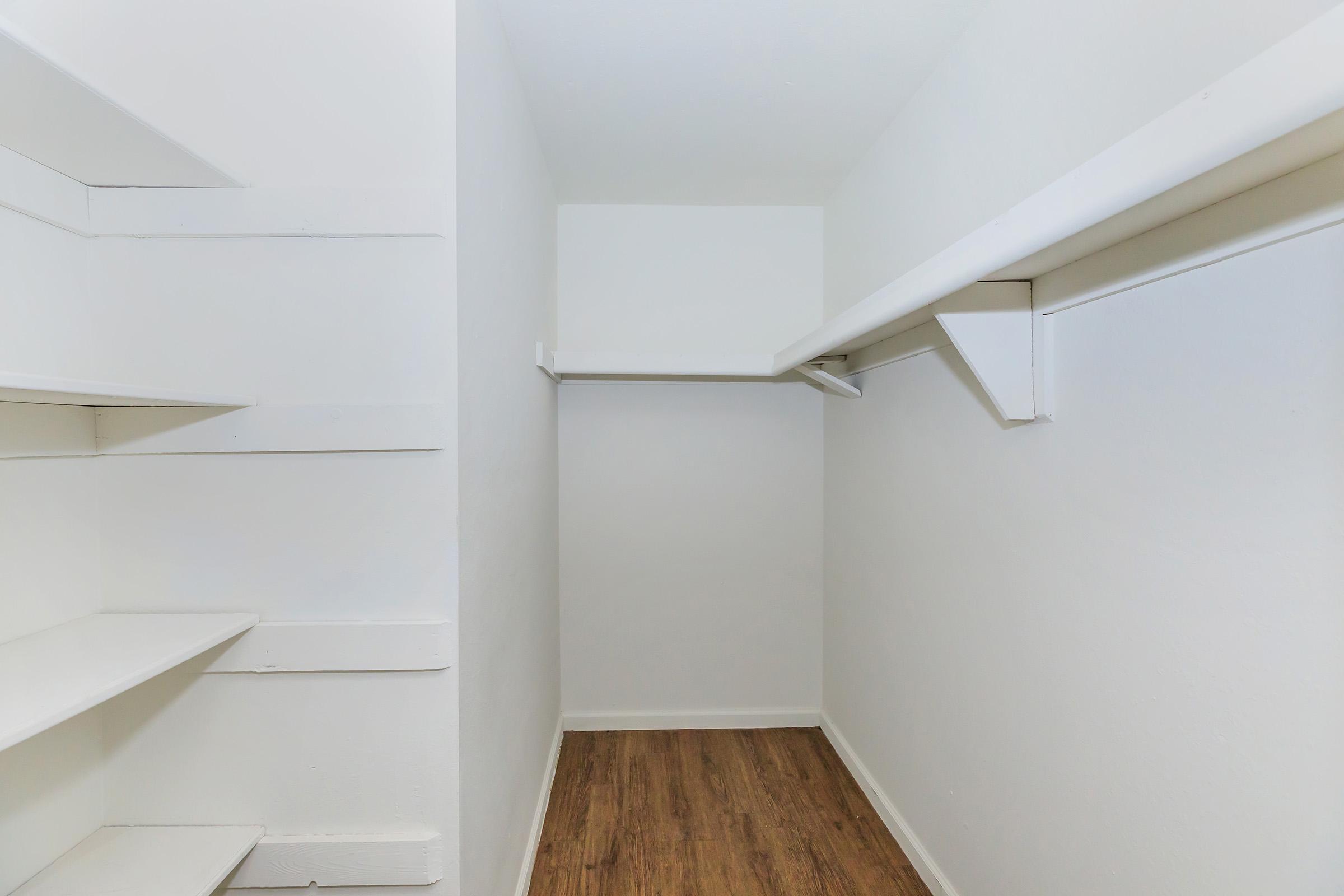 A small, empty closet with white walls and a wooden floor. There are two shelves on the left side, and a hanging rod along the far wall. The space is well-lit and appears clean and tidy.