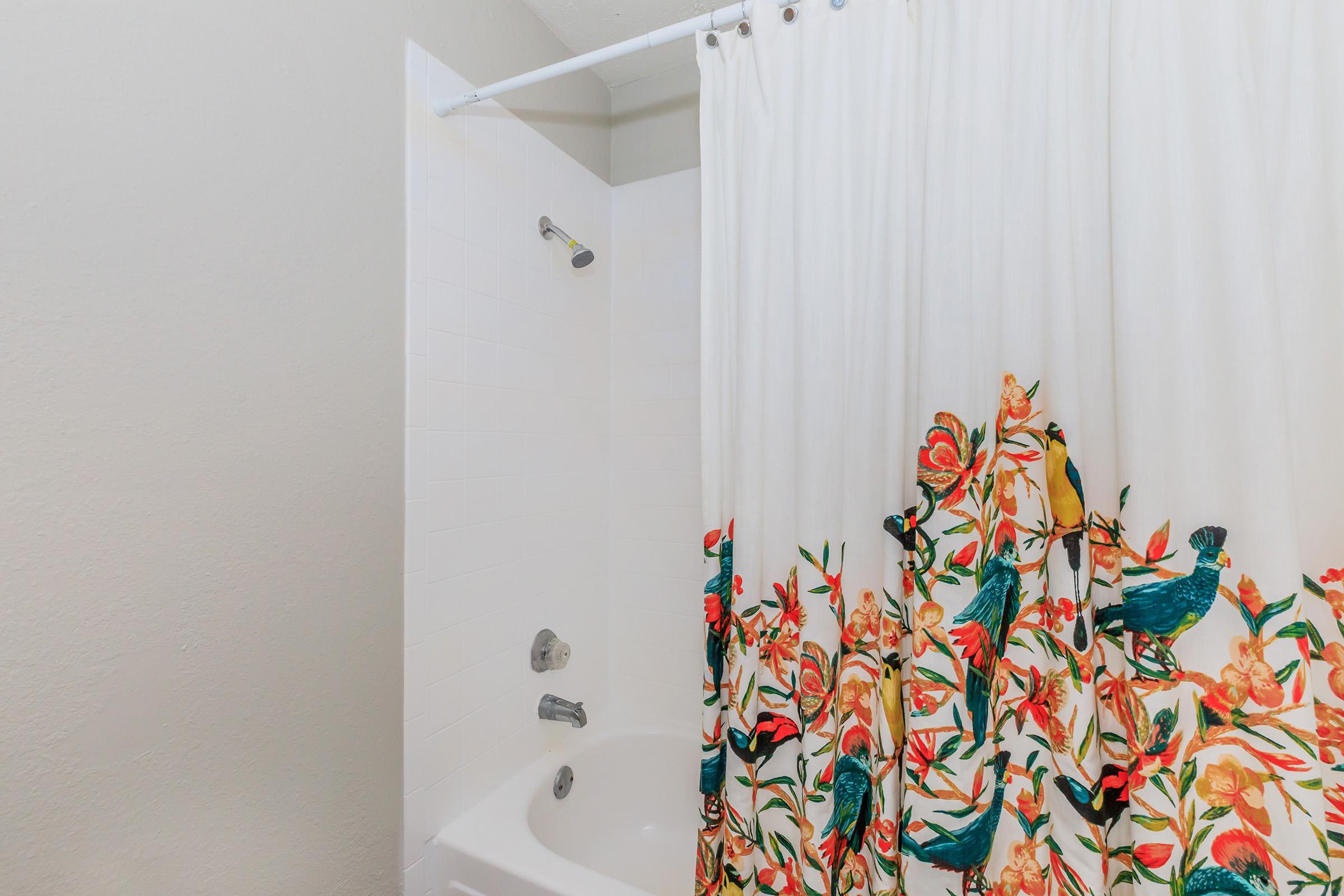 A bathroom scene featuring a white bathtub with a colorful floral shower curtain in shades of red, orange, and green. The walls are painted a light neutral color, and there is a showerhead installed above the tub. The overall atmosphere is bright and inviting.