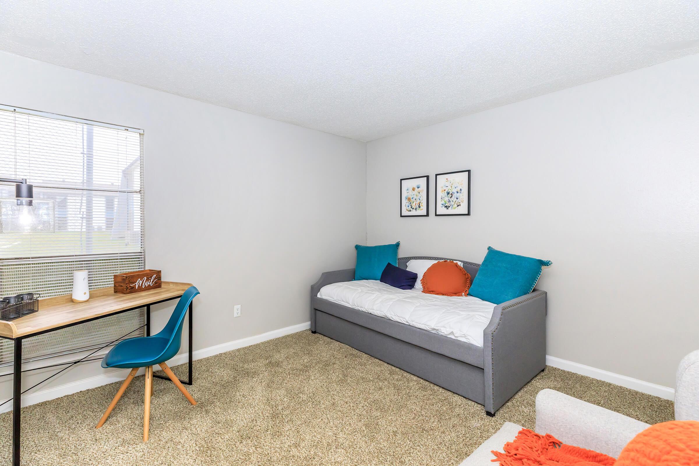 A cozy and minimalist room featuring a gray daybed with colorful pillows, a small desk with a blue chair, and two framed floral prints on the wall. Natural light flows in from a window adorned with blinds, and the floor is covered with soft carpet, creating a welcoming atmosphere.