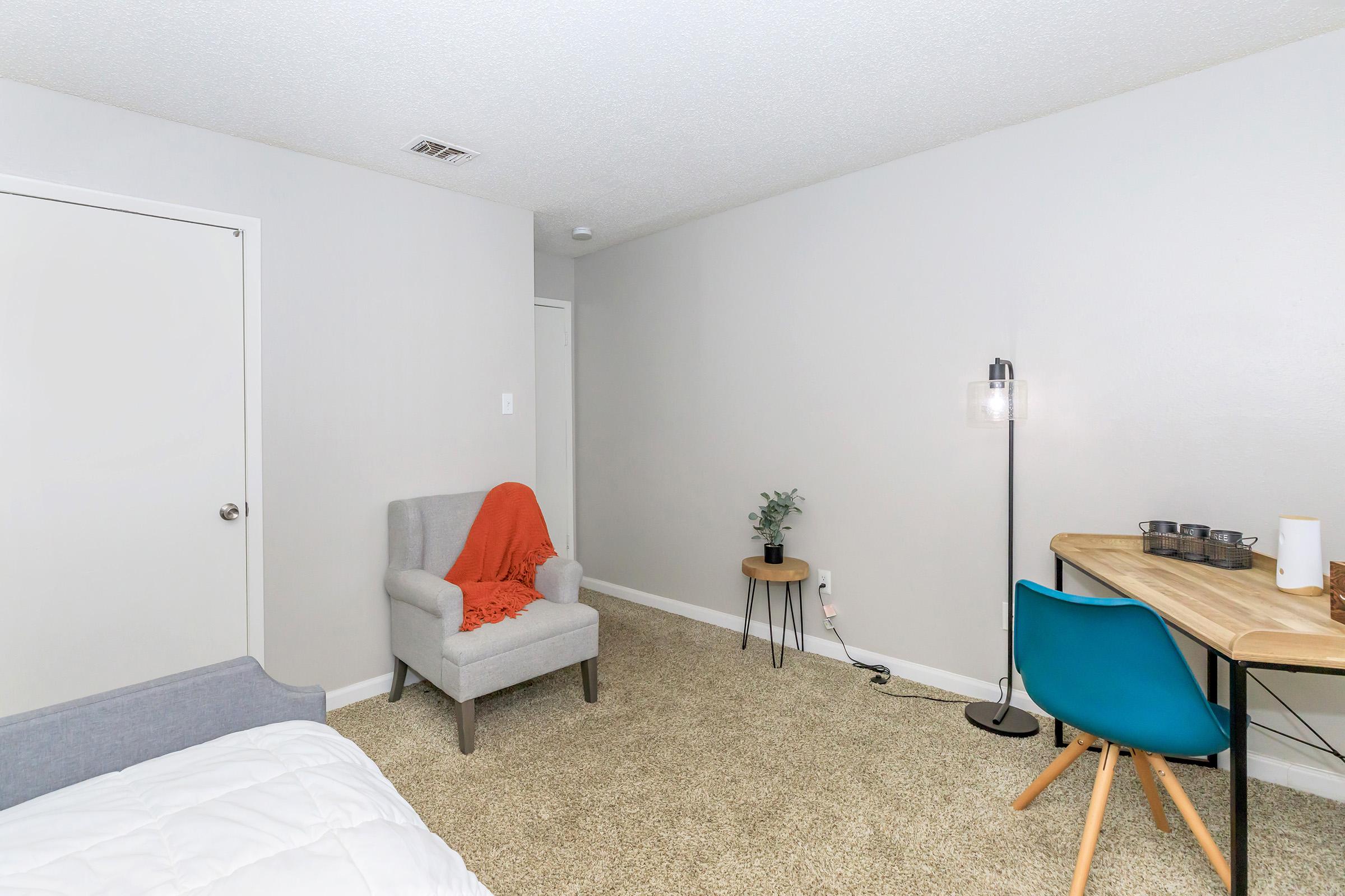 A cozy, minimalistic bedroom featuring a gray armchair with a red throw blanket, a small side table with a plant, and a wooden desk with chairs. The walls are painted light gray, and the floor is covered with carpet. Natural light illuminates the space, creating a warm atmosphere.
