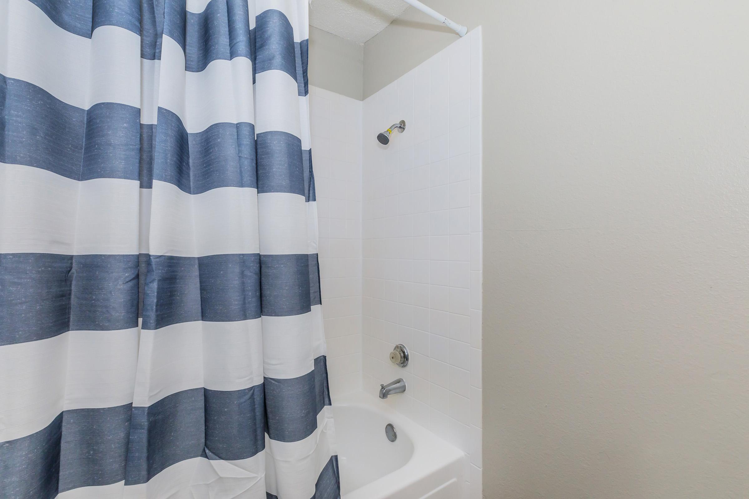 A bathroom shower area featuring a white tub, a white tiled wall, and a shower curtain with blue and white stripes. The showerhead is mounted on the wall, and the overall space is clean and well-lit.