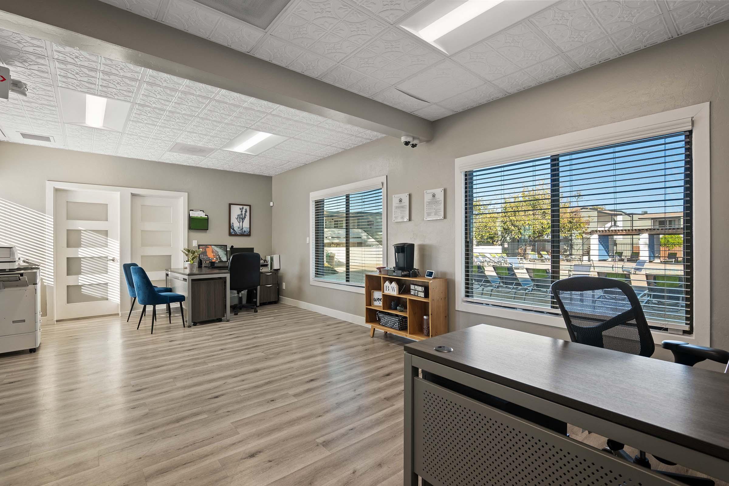 A brightly lit office space featuring two desks, an office chair, and cabinets. Large windows provide natural light and a view of the outdoors. The decor is modern with wood-like flooring and an organized, professional atmosphere.