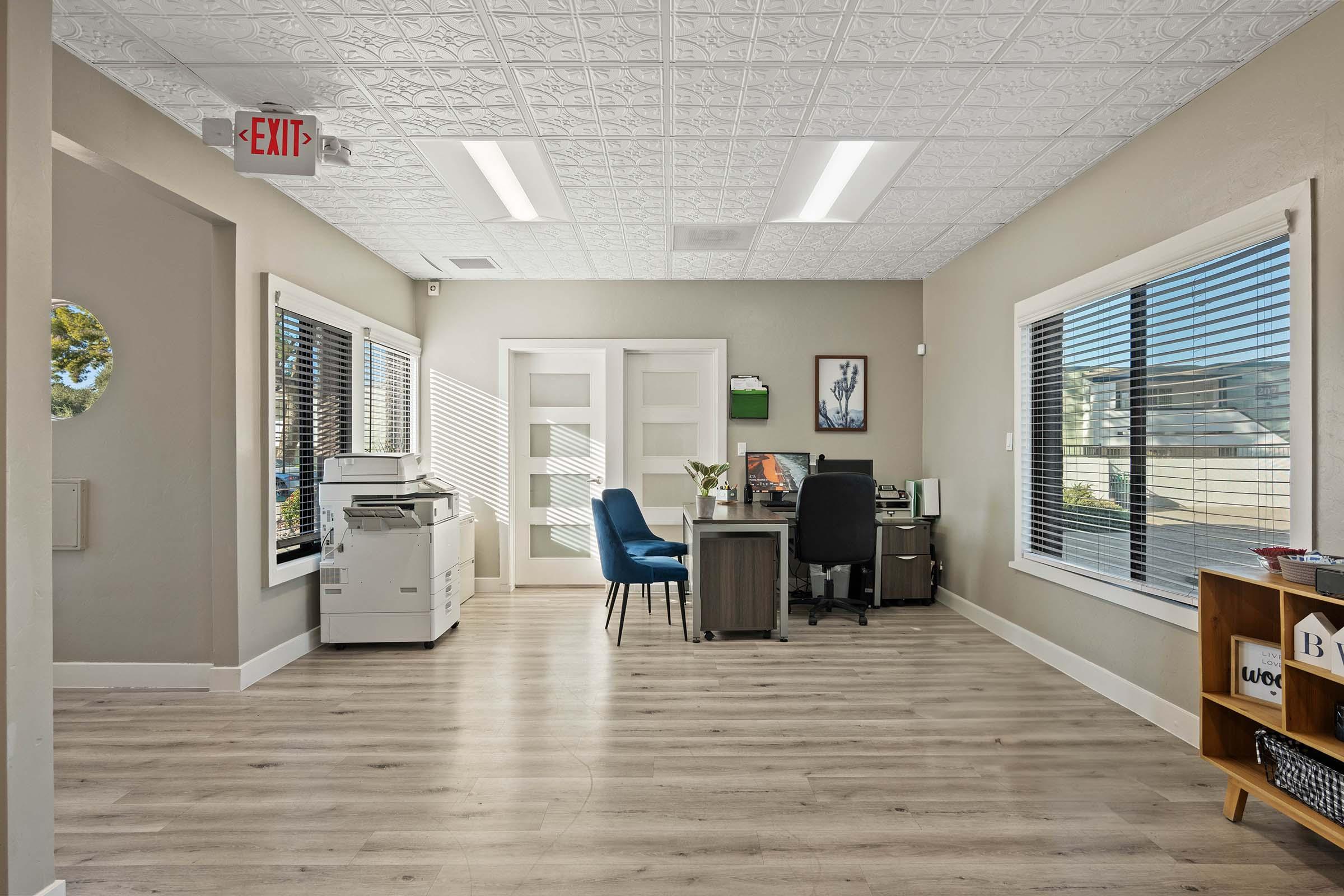 A modern office space featuring a desk with a chair, a printer, and large windows allowing natural light. The walls are painted in a neutral color, and there’s a small plant and artwork on display. The flooring is wood-like, and there's a sign indicating an exit.