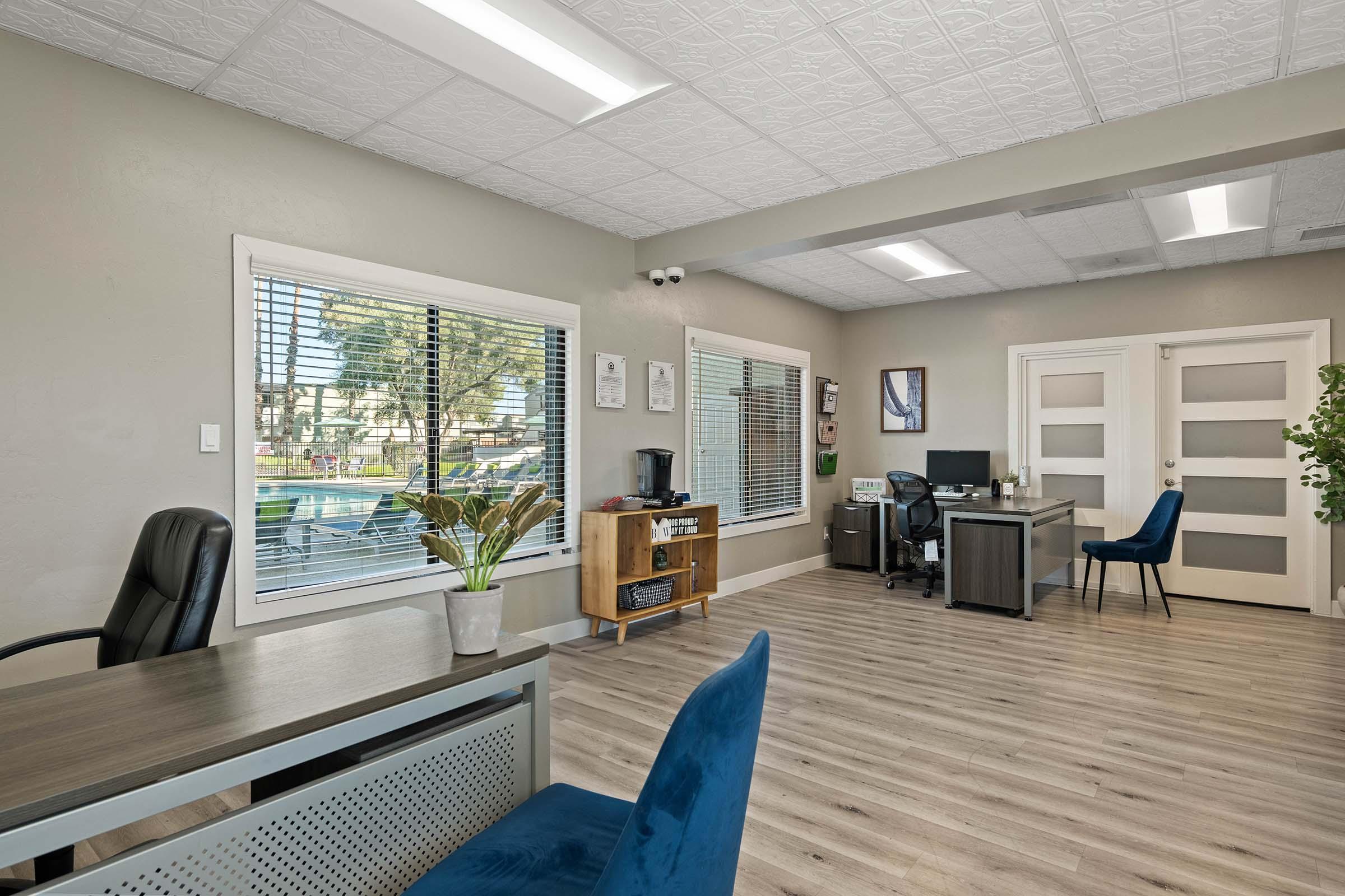 Interior of a modern office space featuring a desk, a blue chair, and a window with blinds. There is a plant on the desk, a coffee machine, and another desk with a computer setup visible. The flooring is wooden, and there are frosted glass doors leading to another area. Natural light illuminates the room.
