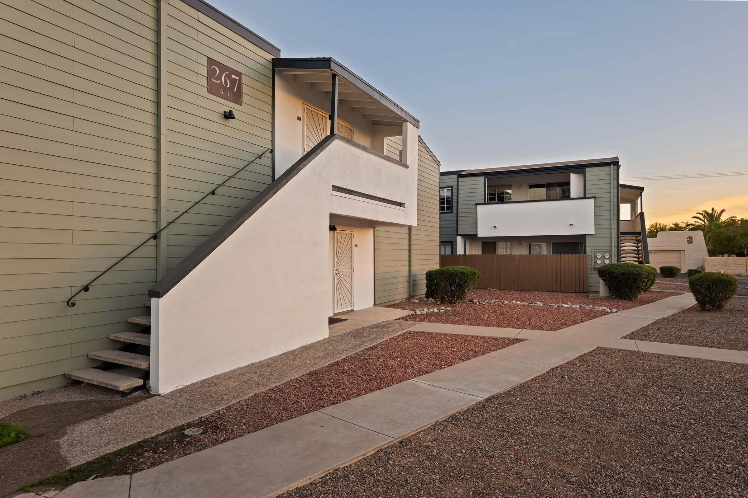 A view of a modern apartment complex featuring two buildings. One building shows a staircase leading to the upper level with a door, while the other has a white facade and is set back from the pathway. The landscaped area includes gravel, small shrubs, and a clear sky in the background, indicating early evening.