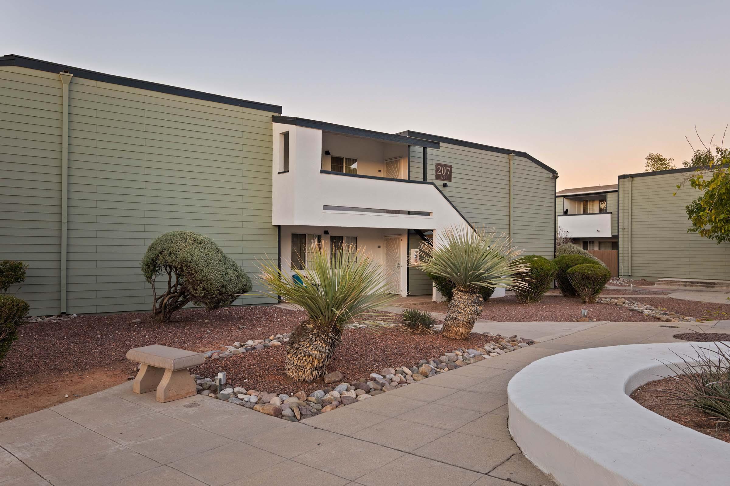 A modern apartment complex with green siding features a central courtyard. In the foreground, a curved pathway leads to a stone bench surrounded by desert landscaping, including palm trees and gravel. The building has two levels with a prominently displayed number above the entrance, set against a soft evening sky.