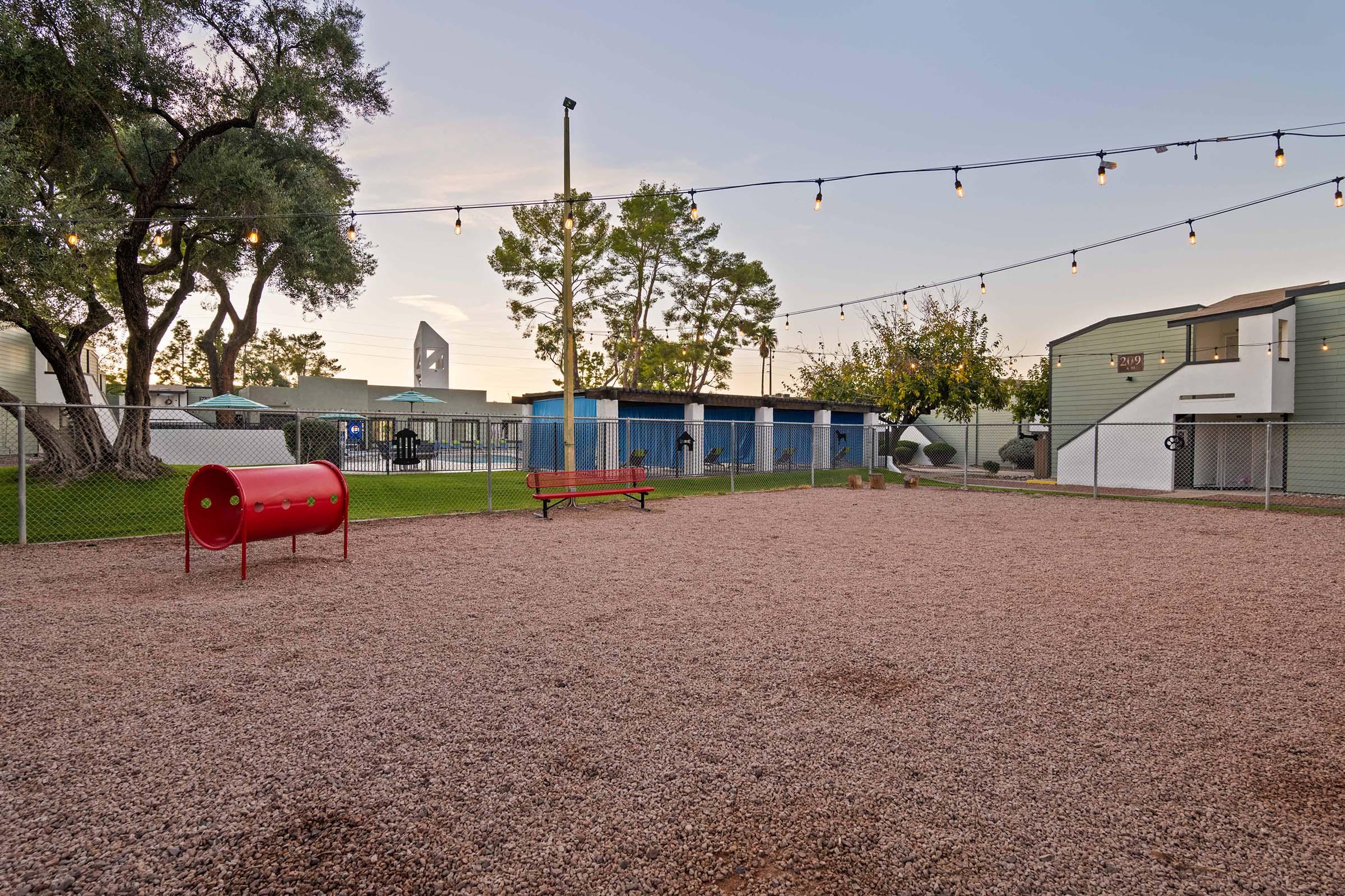 A fenced playground area featuring a red cylindrical object, a wooden bench, and gravel ground. Surrounding the space are trees, string lights overhead, and several buildings in the background, creating a welcoming and recreational atmosphere.